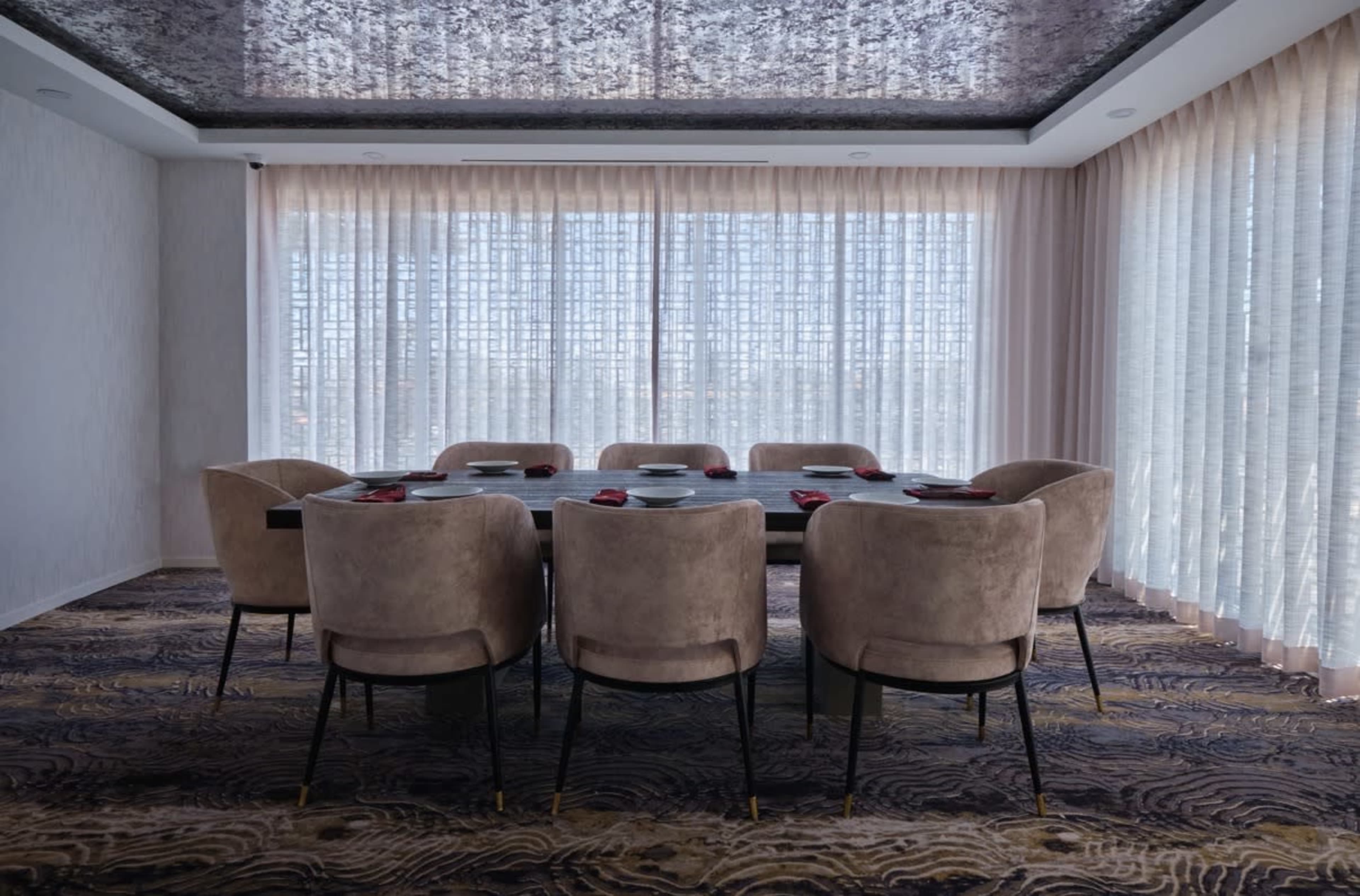 A long dining table with eight upholstered chairs is set in a well-lit room featuring large, sheer-curtained windows.