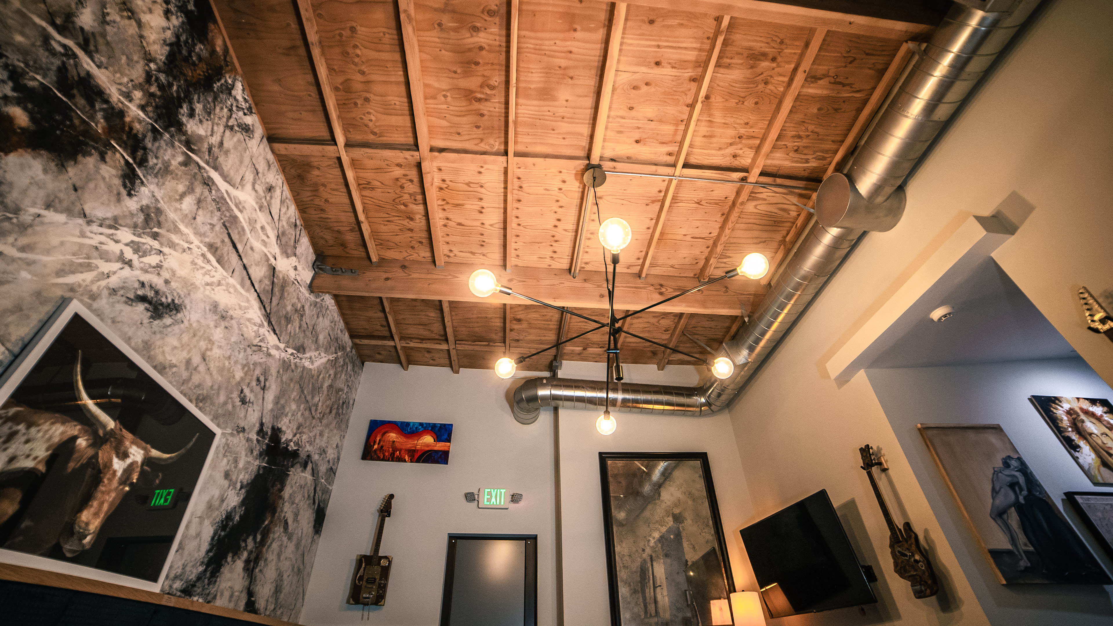 The image shows a modern interior with a wooden ceiling, industrial-style lighting, and various artworks on the walls.