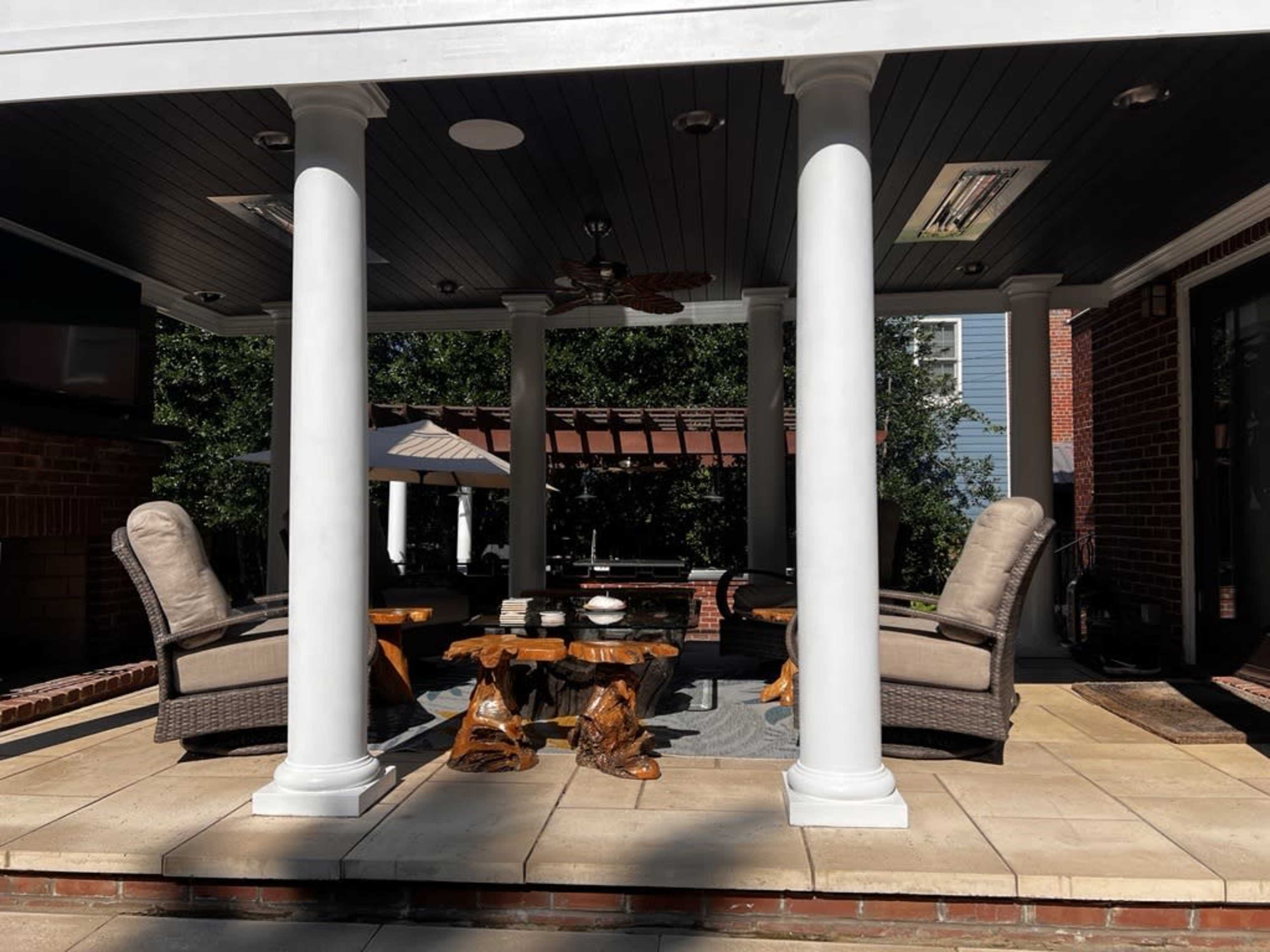 A covered patio area with two chairs, a wooden table, and four white columns, surrounded by greenery.