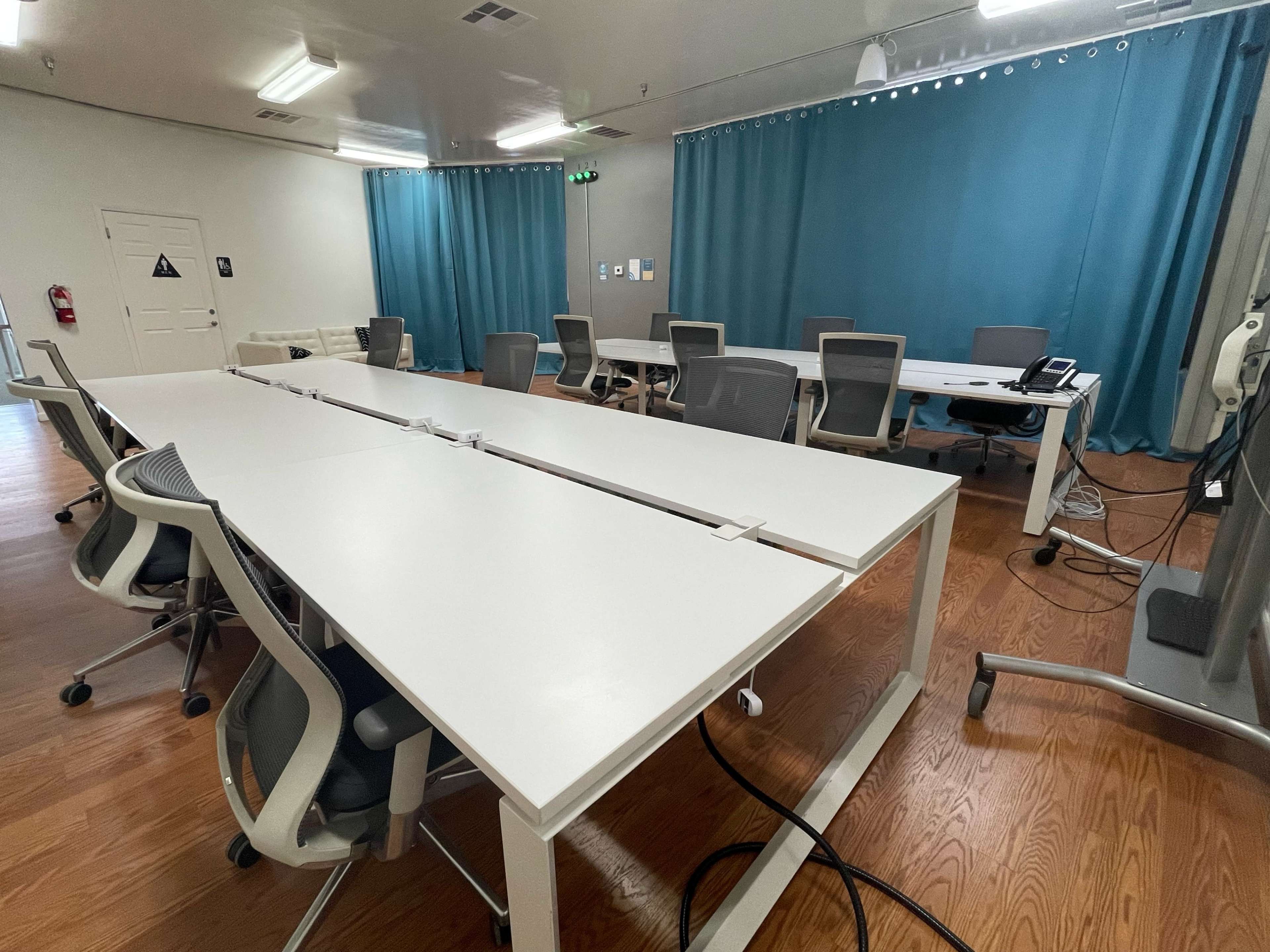A modern conference room with multiple white tables and ergonomic chairs, separated by a blue curtain in the background.