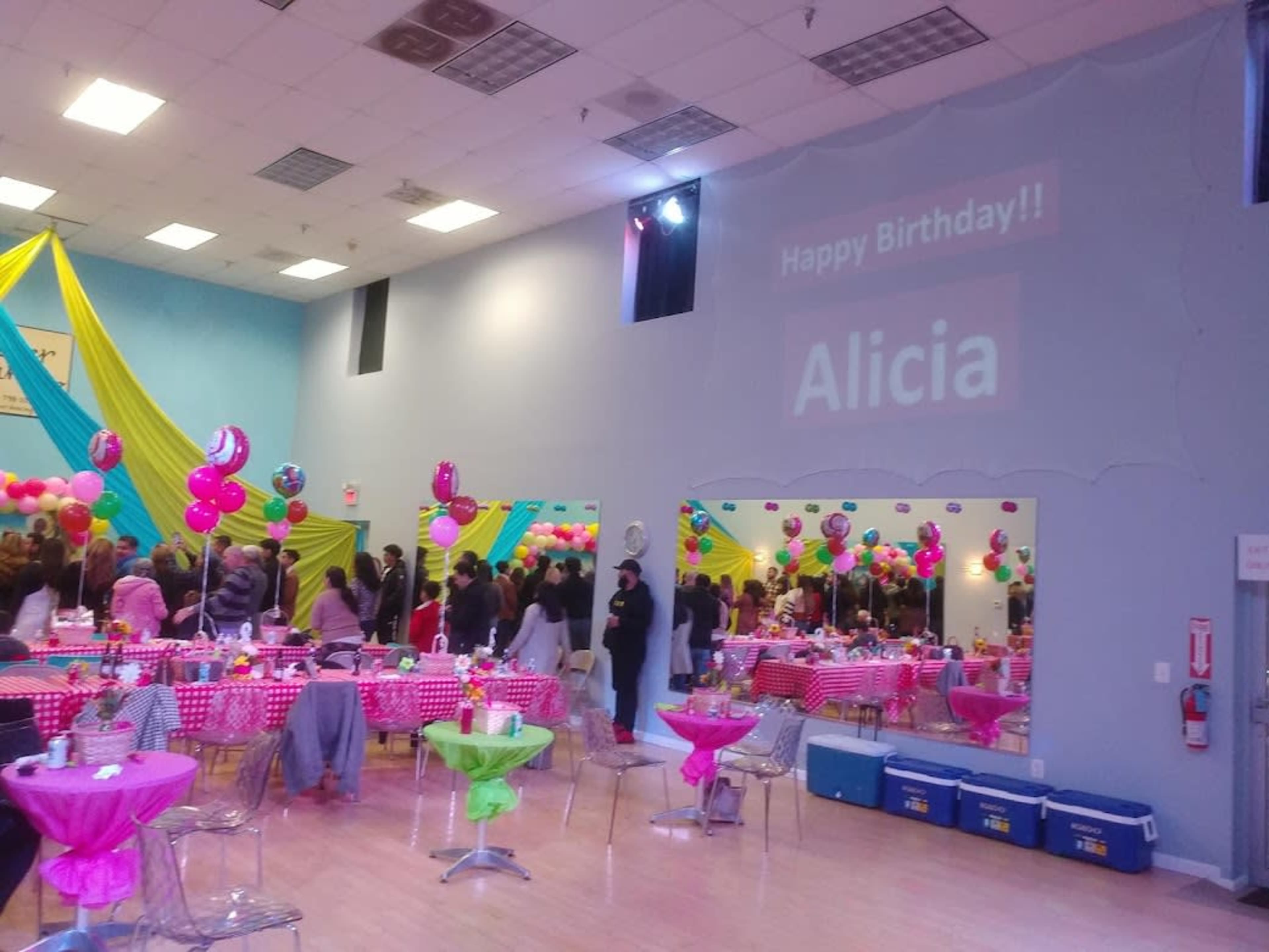 A large birthday party is set up in a colorful room with tables decorated in pink and green, and a wall display projecting "Happy Birthday!! Alicia."