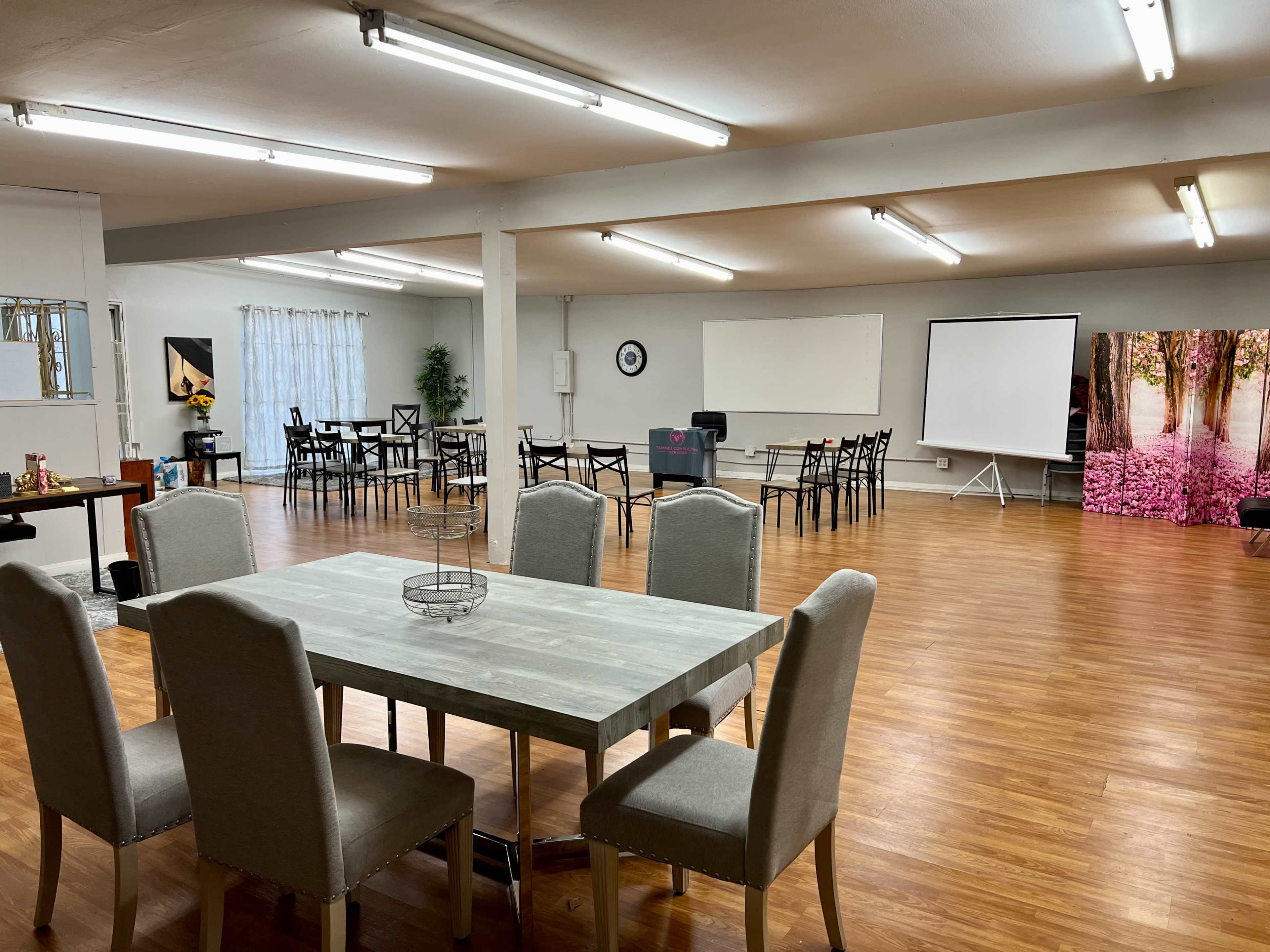 A spacious room with a dining table and chairs in the foreground, and a seating area arranged with additional chairs and a projector screen in the background.