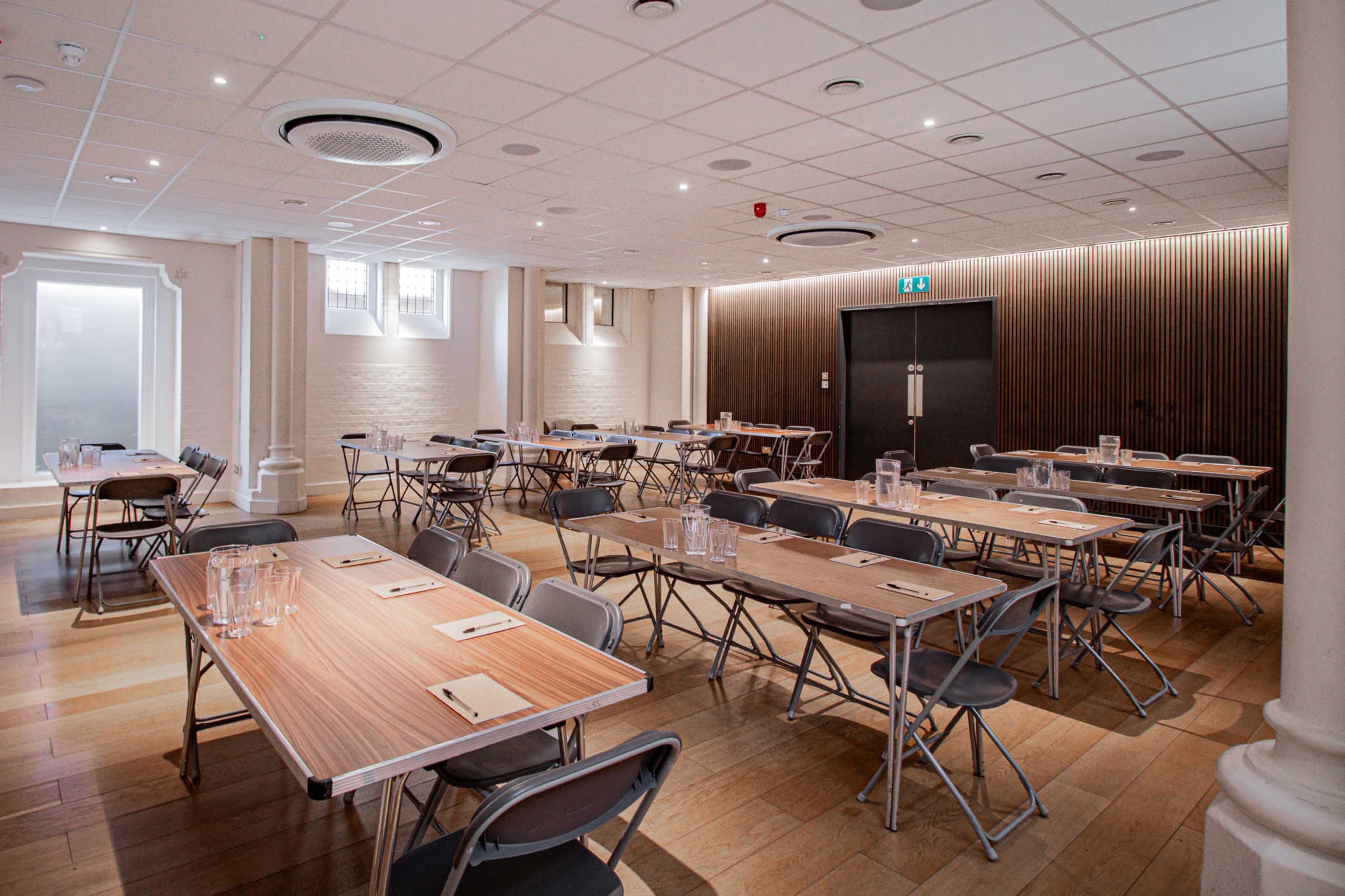 The image shows a spacious meeting room with multiple wooden tables set up in rows, accompanied by folding chairs and glass water pitchers on each table.