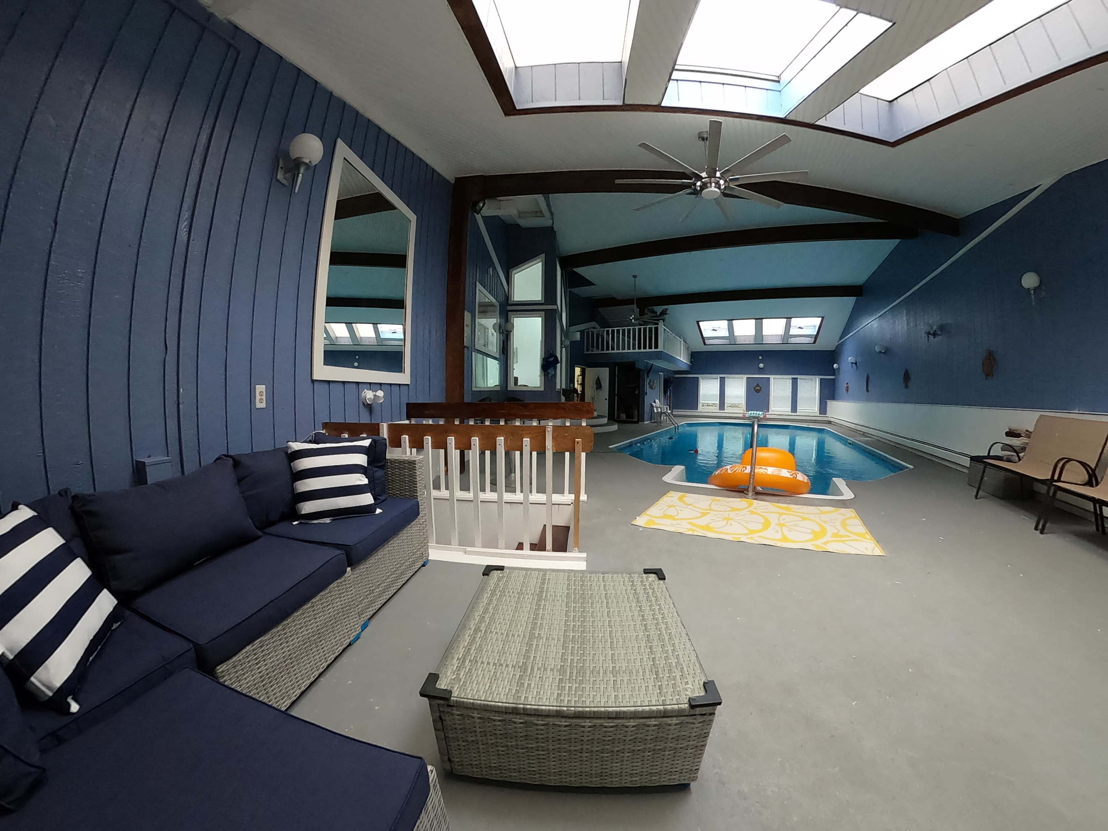 A blue-walled indoor pool area with a seating arrangement, a coffee table, and pool features including a float and skylights.