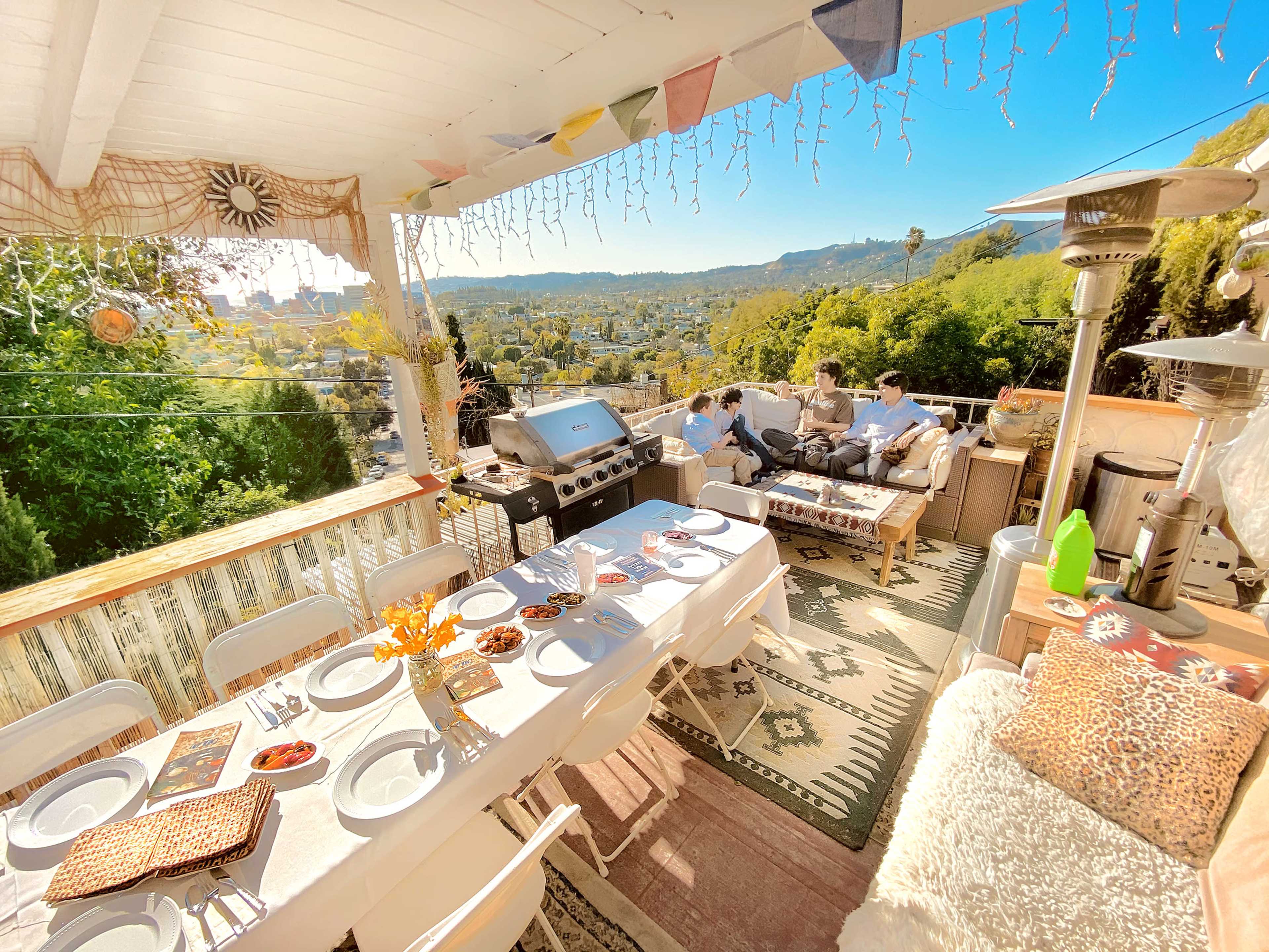 A spacious outdoor dining area featuring a long table set for a meal, a grill, and a view of a hillside landscape.