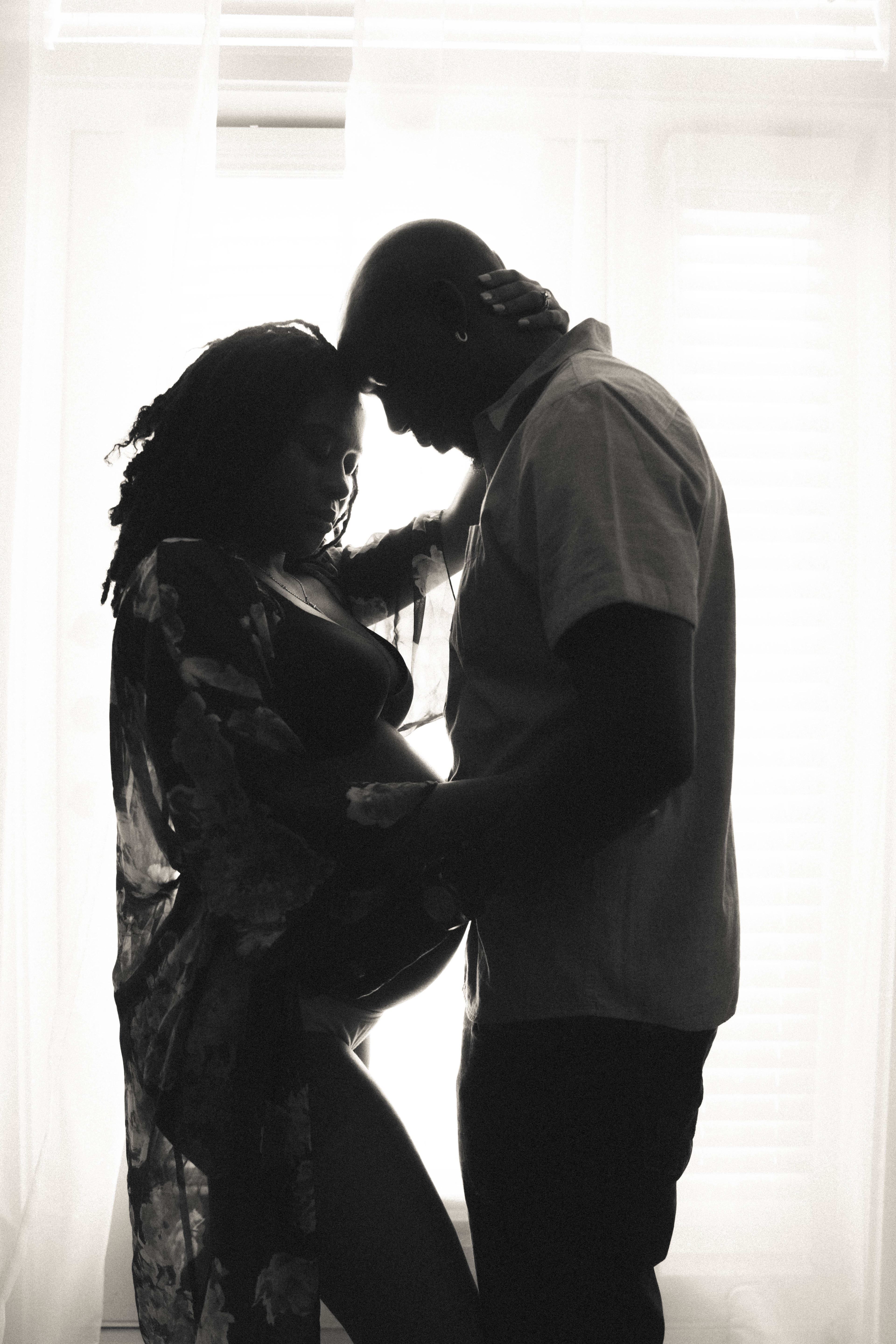 A pregnant woman and her partner posing for a black and white maternity photoshoot in front of a window.