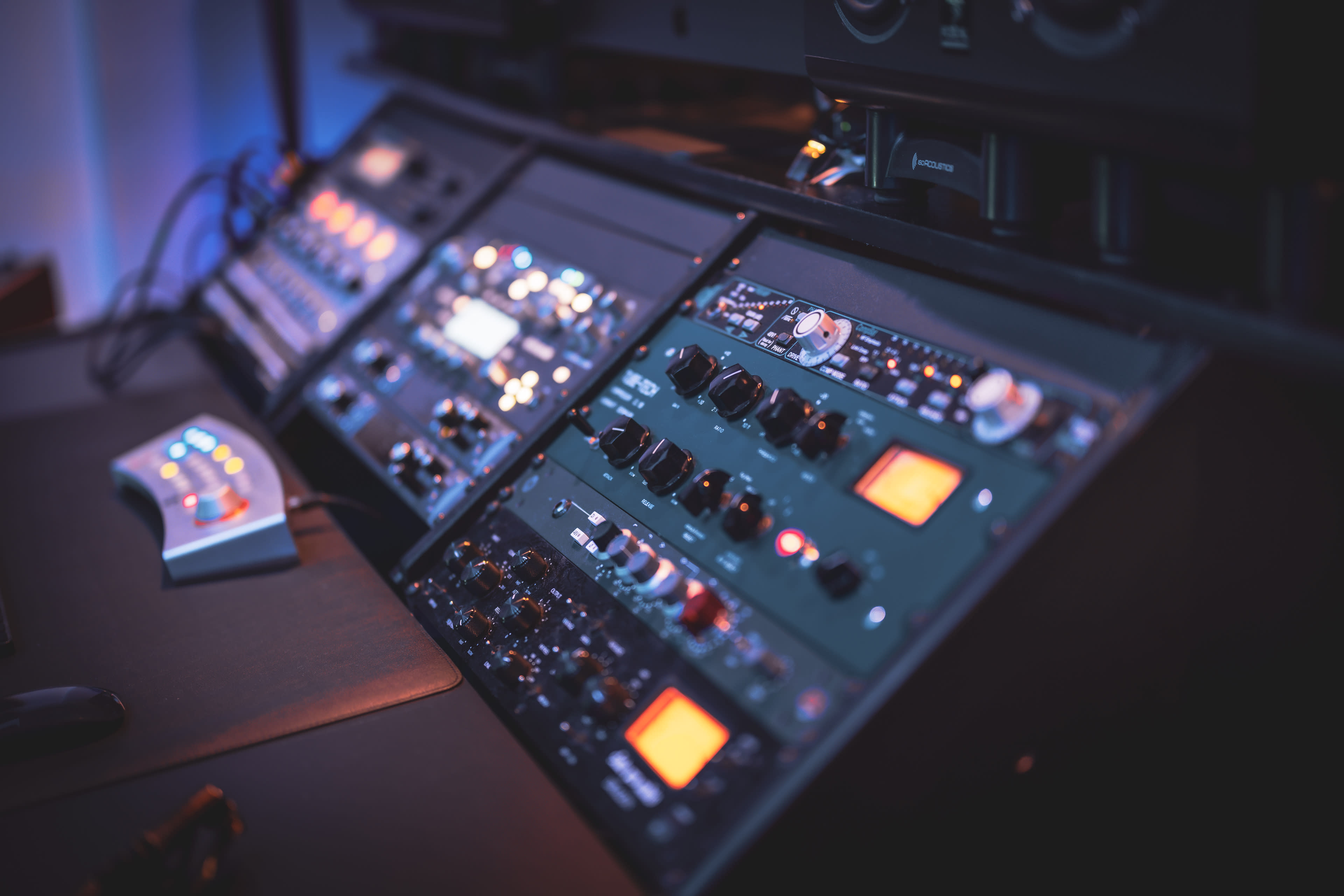 The image shows a close-up of audio mixing equipment with various knobs, buttons, and displays illuminated in dim lighting.