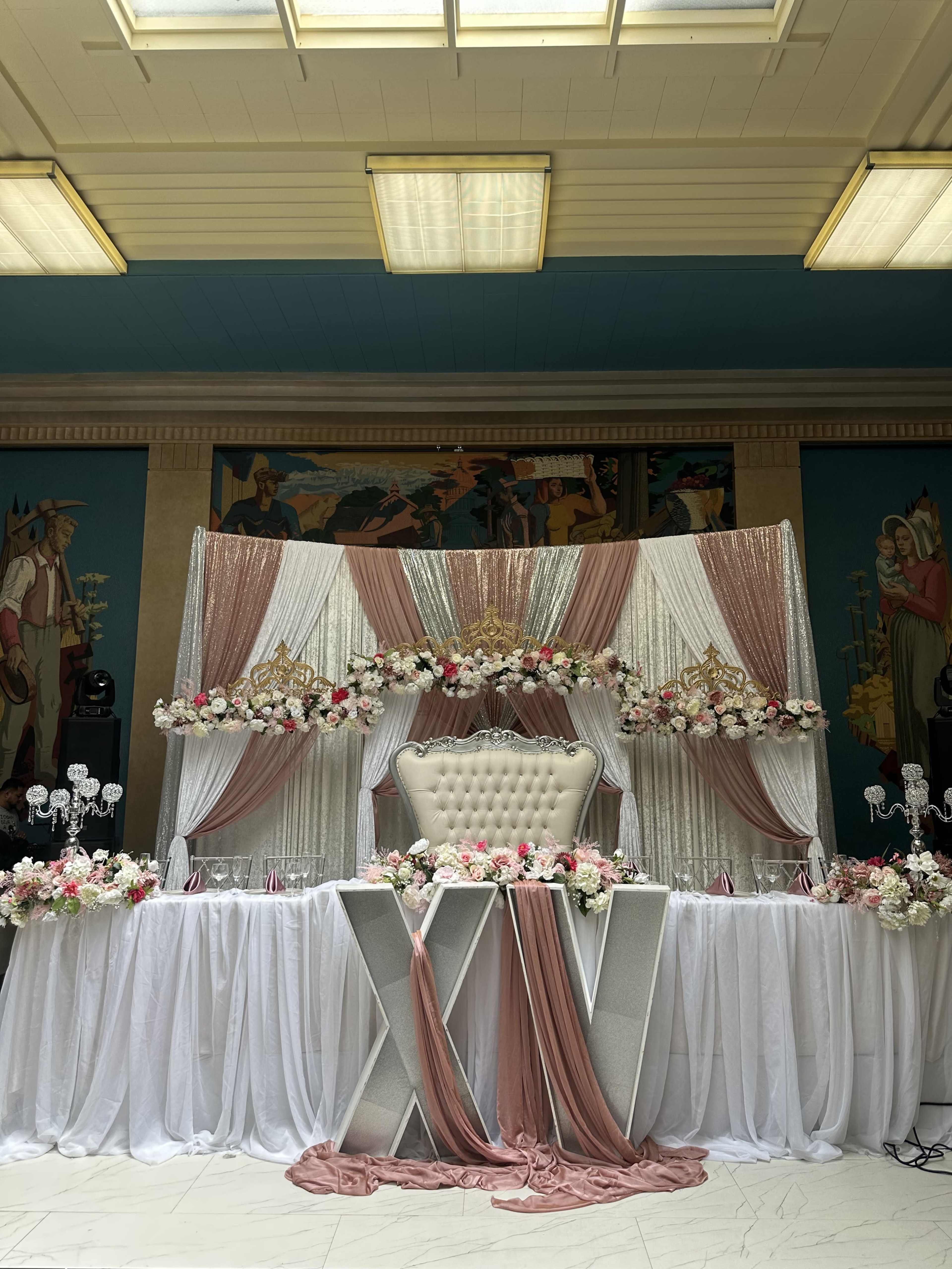 The image shows a decorated event space with a white and pink themed backdrop, featuring floral arrangements and a central lavish white throne.