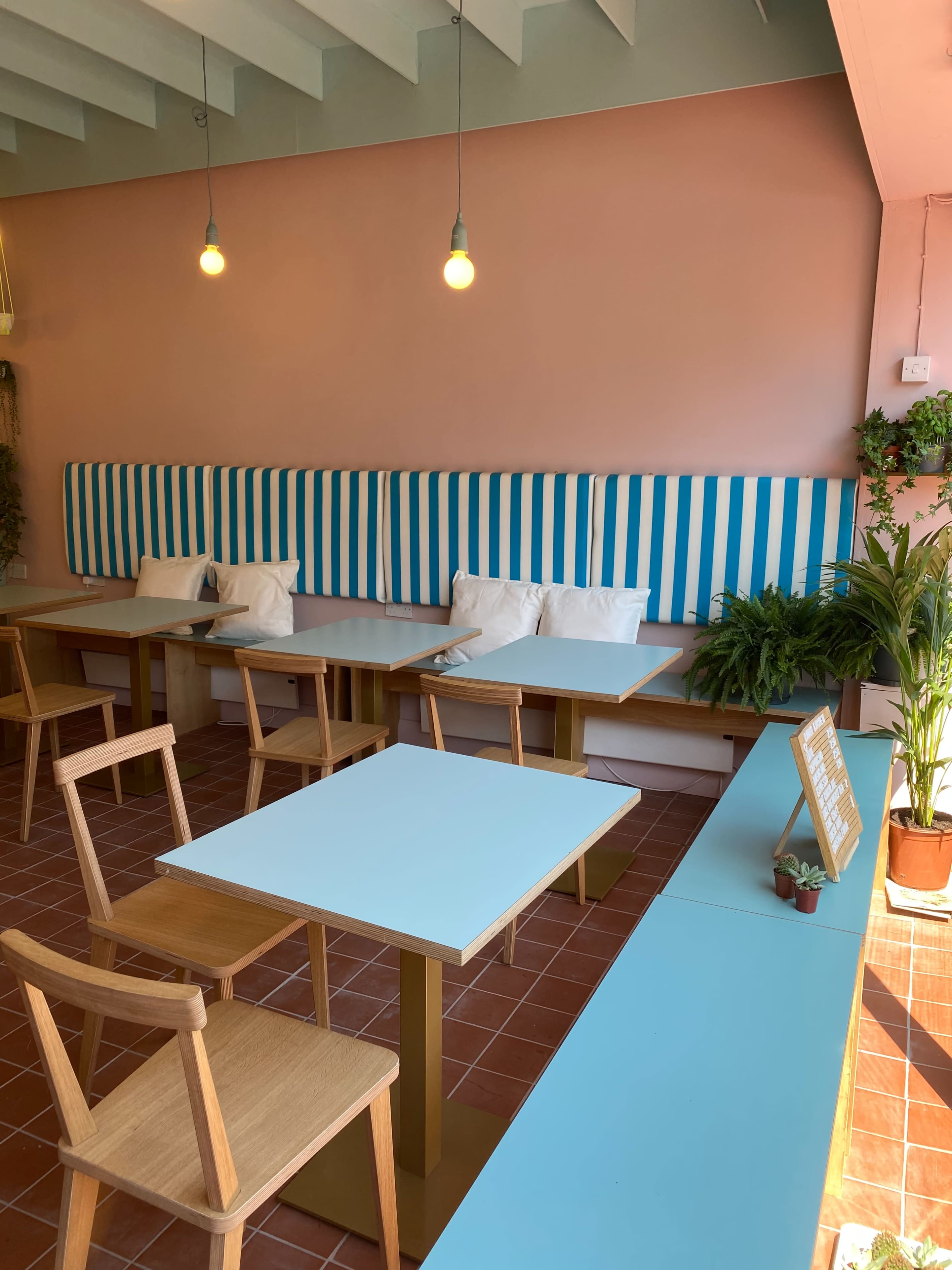 A brightly lit café interior with striped seating along the wall, wooden tables, and plants along the edges.