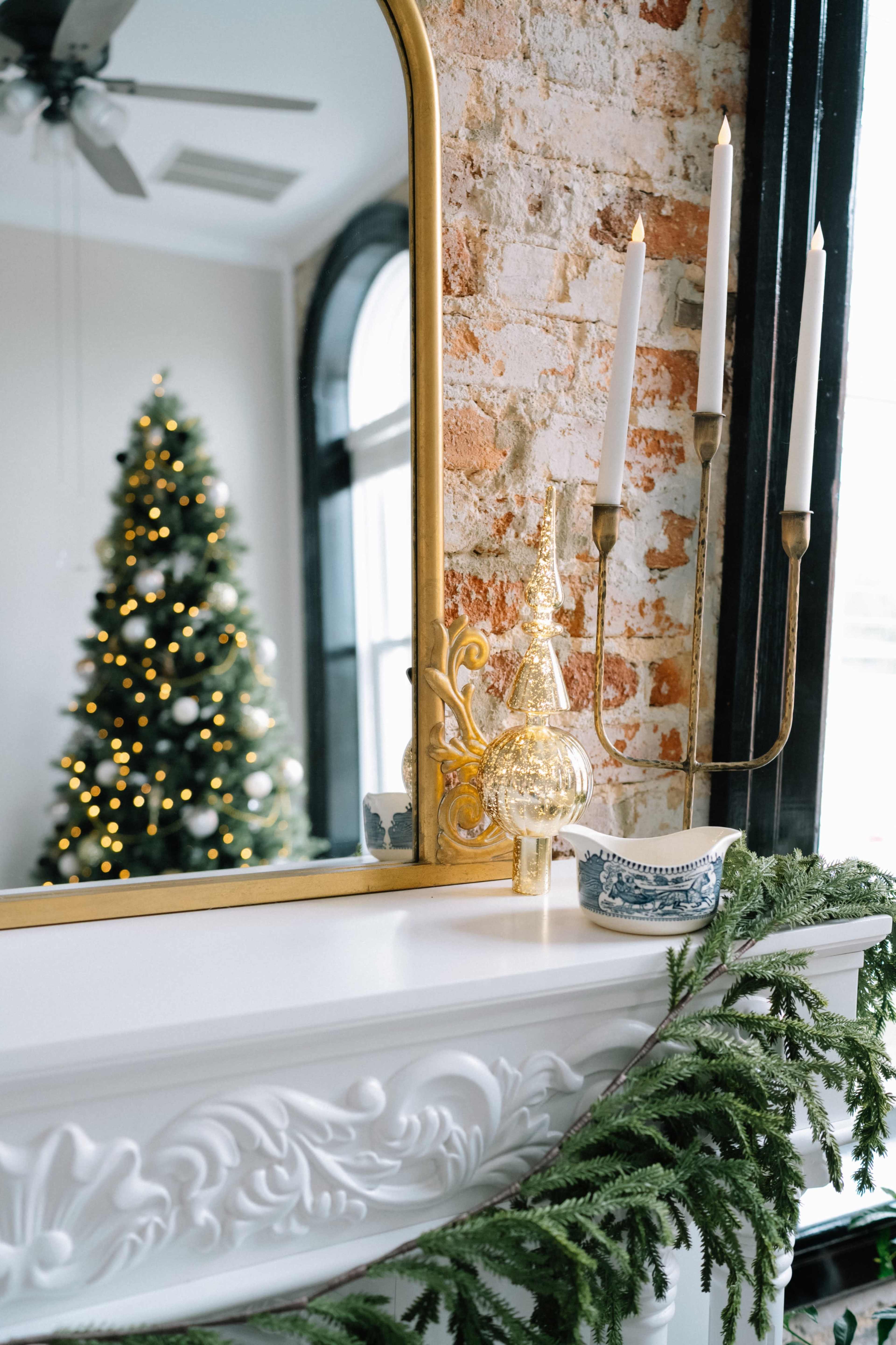 A decorated mantle features a gold mirror, a decorative glass tree, three lit candles, and a garland, with a Christmas tree reflecting in the background.