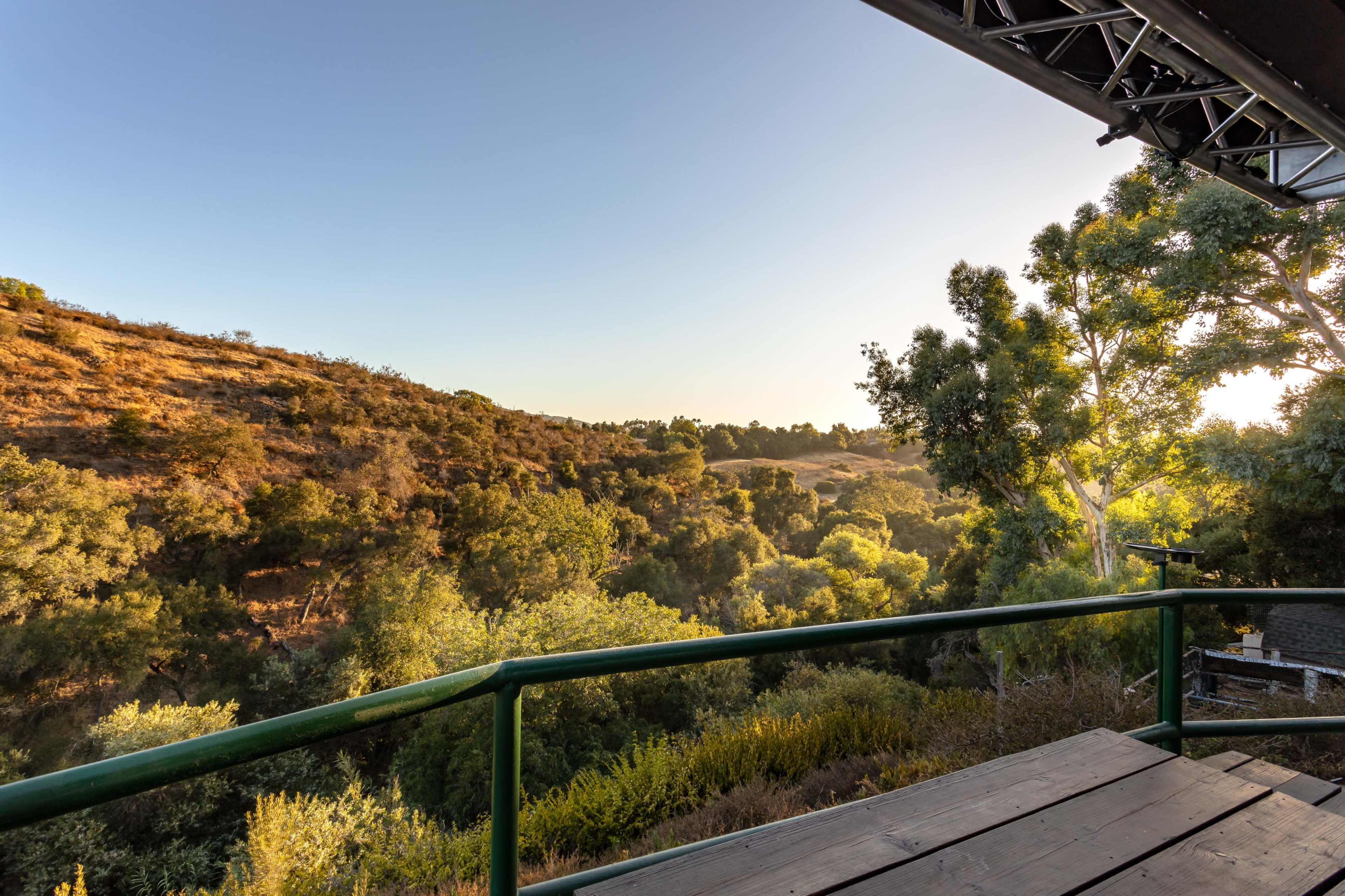 A green balcony railing overlooks a hilly landscape filled with trees under a clear sky.