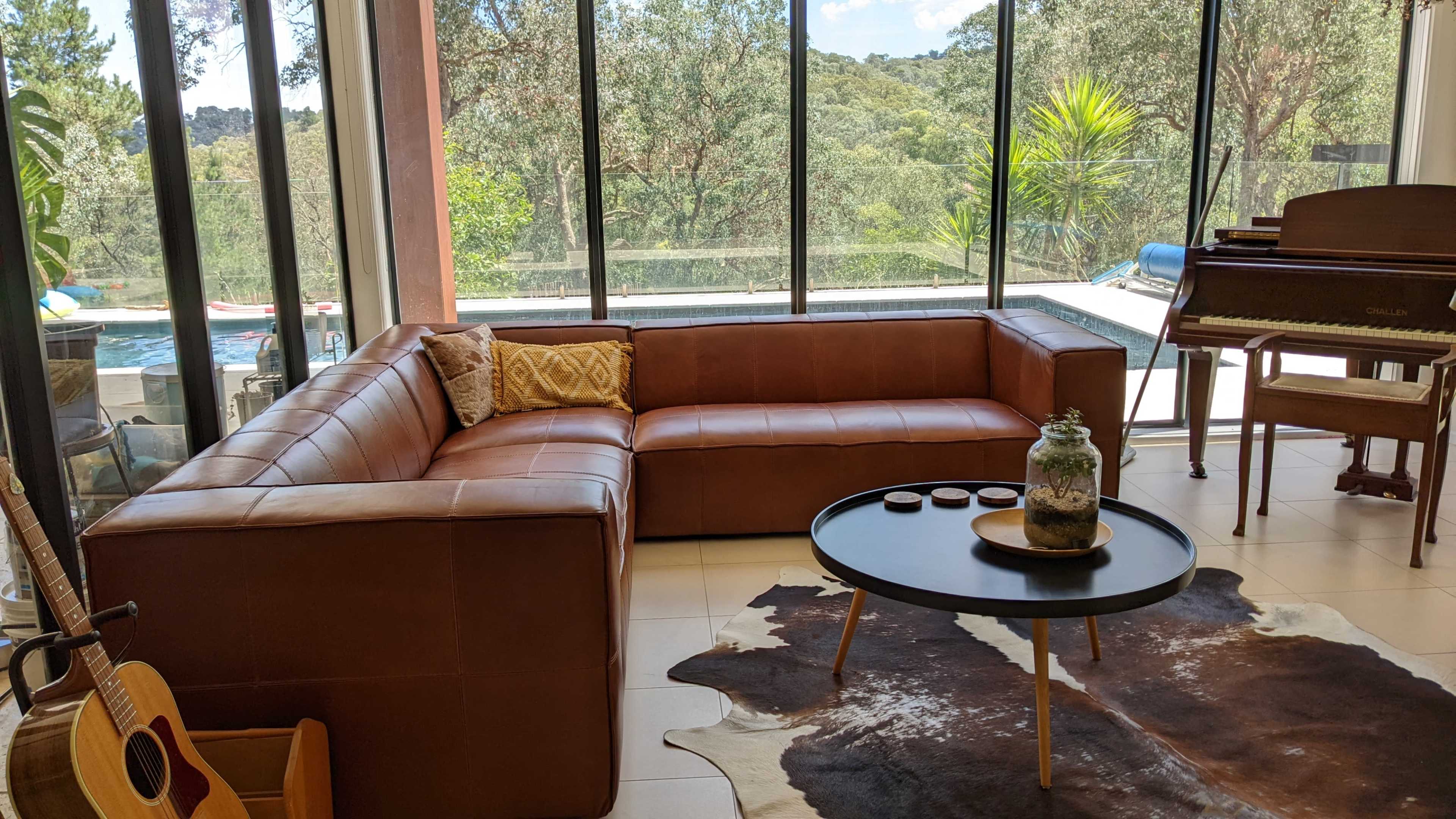 A modern living room features a brown leather sectional sofa, a round coffee table on a cowhide rug, and large windows overlooking a green landscape.