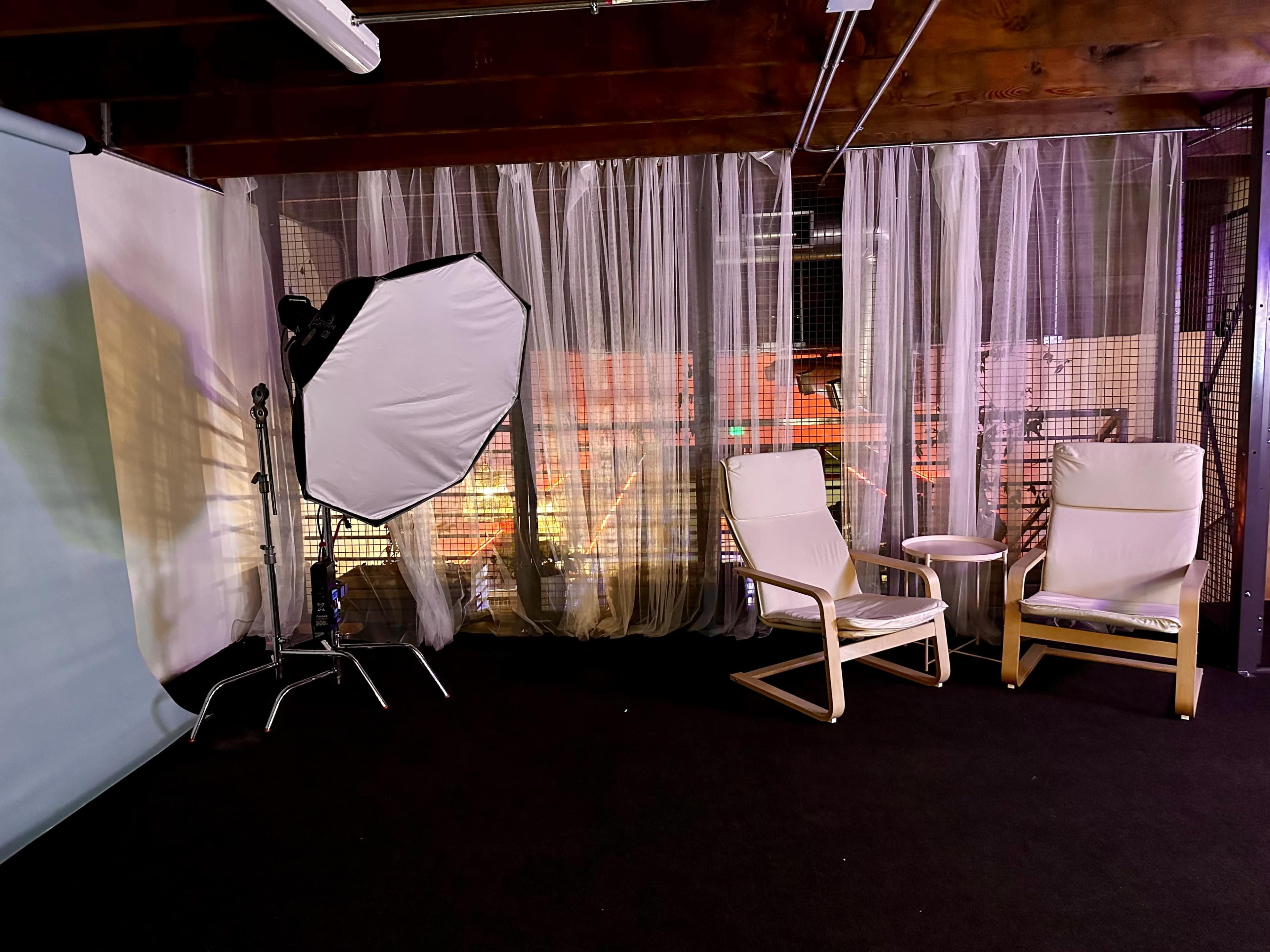 A photography studio with a softbox light, two chairs, and sheer curtains in a dimly lit space.