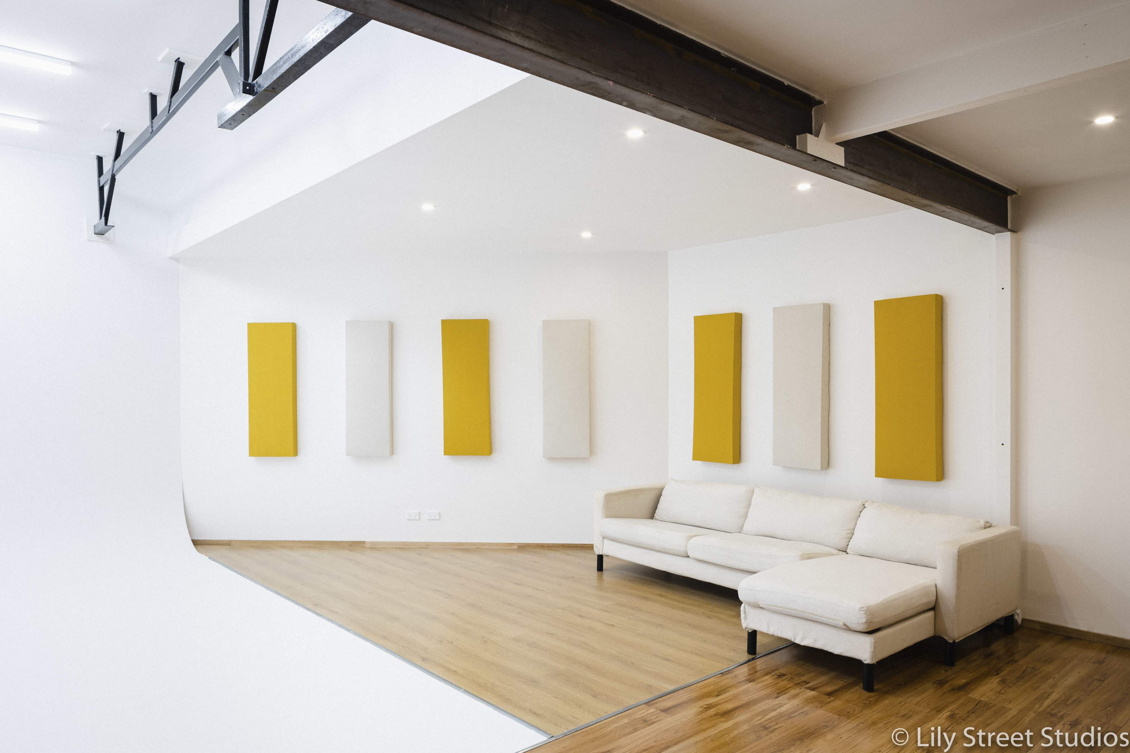 The image shows a modern, bright room with a white curved backdrop, yellow wall panels, and a white sofa against a wooden floor.