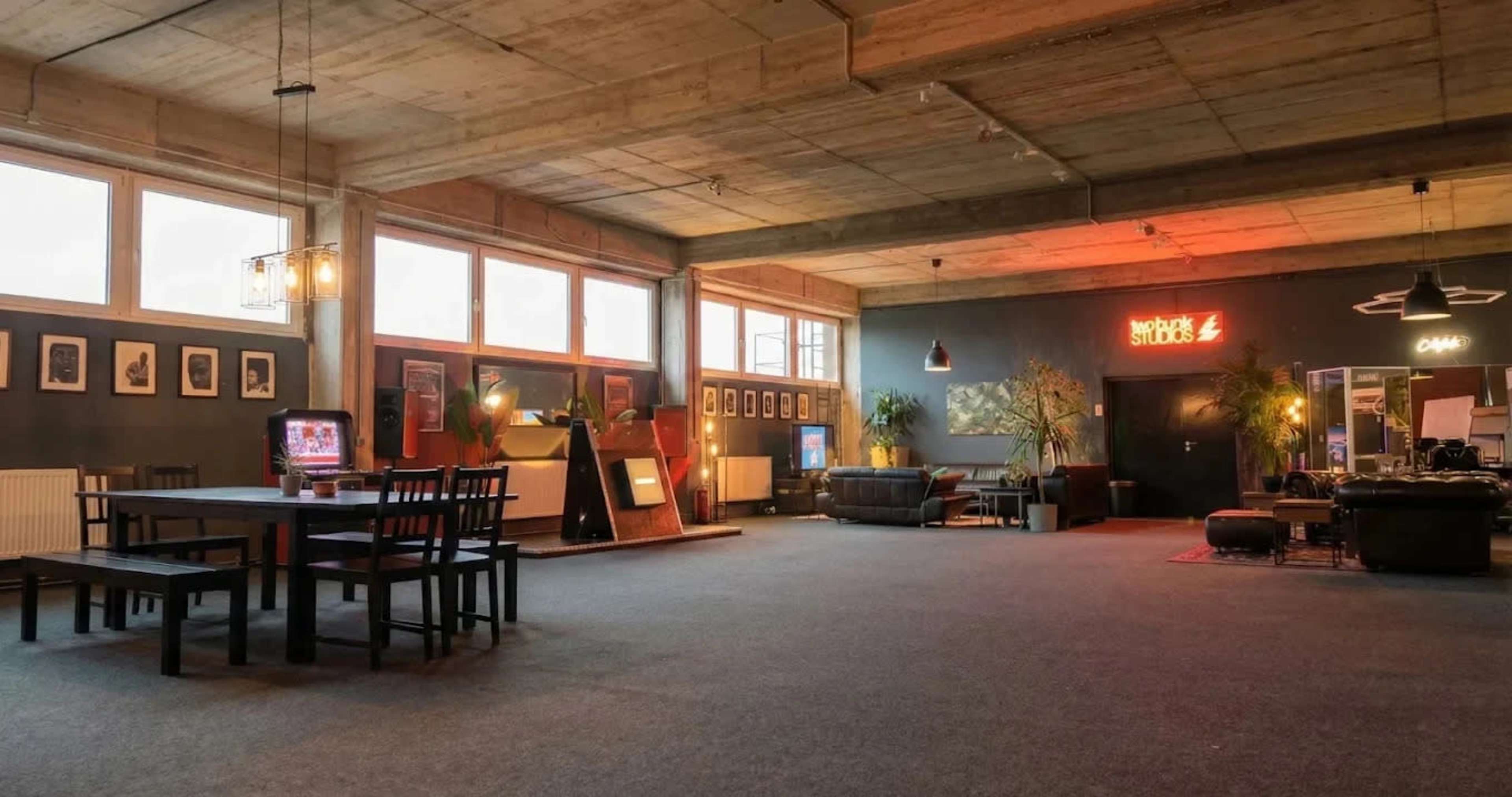 The image depicts an expansive studio space featuring a dining table, seating area, and various decorative elements, including plants and artwork on the walls.