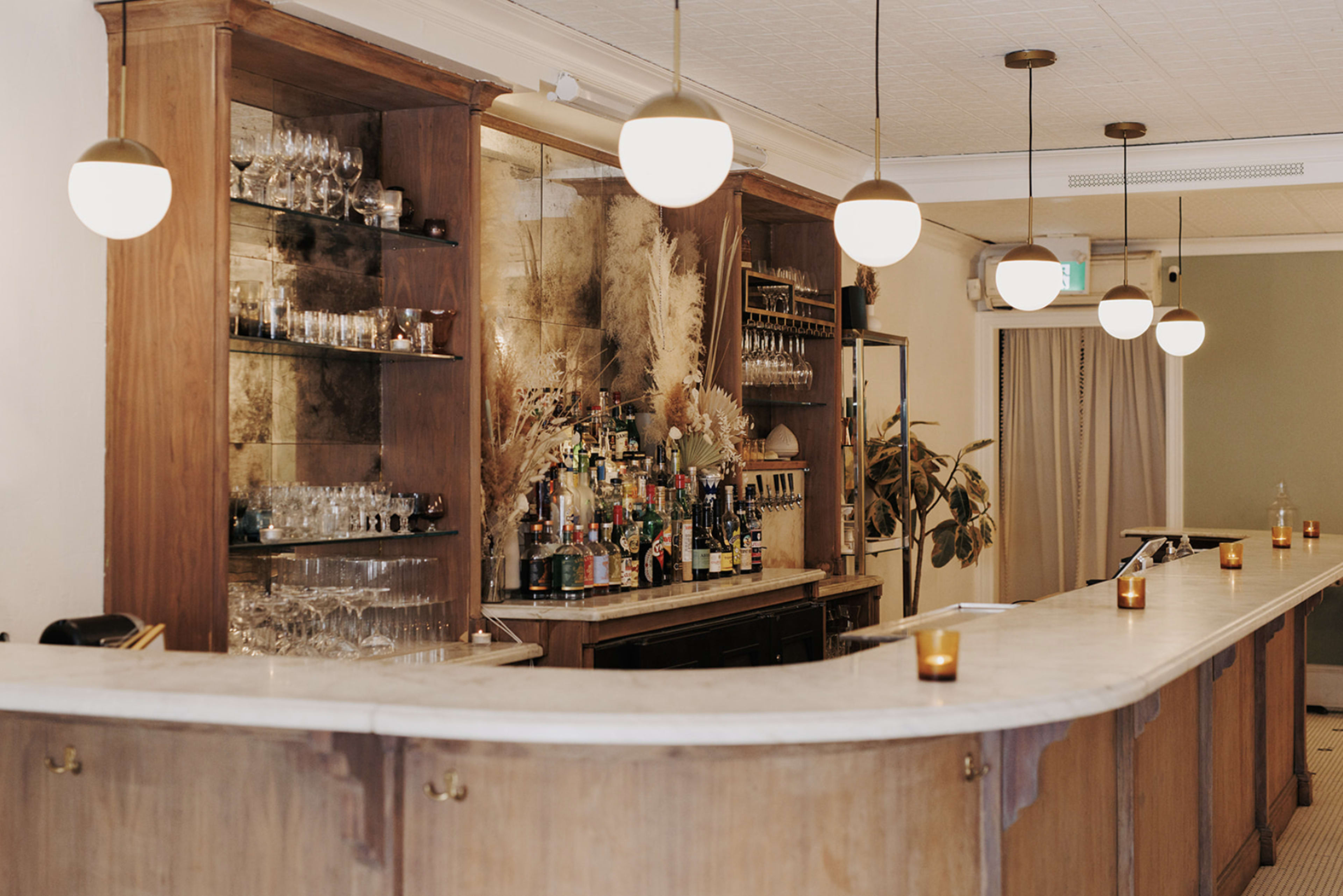 A wooden bar area features a collection of bottles and glasses, with pendant lights hanging above and candles placed along the counter.