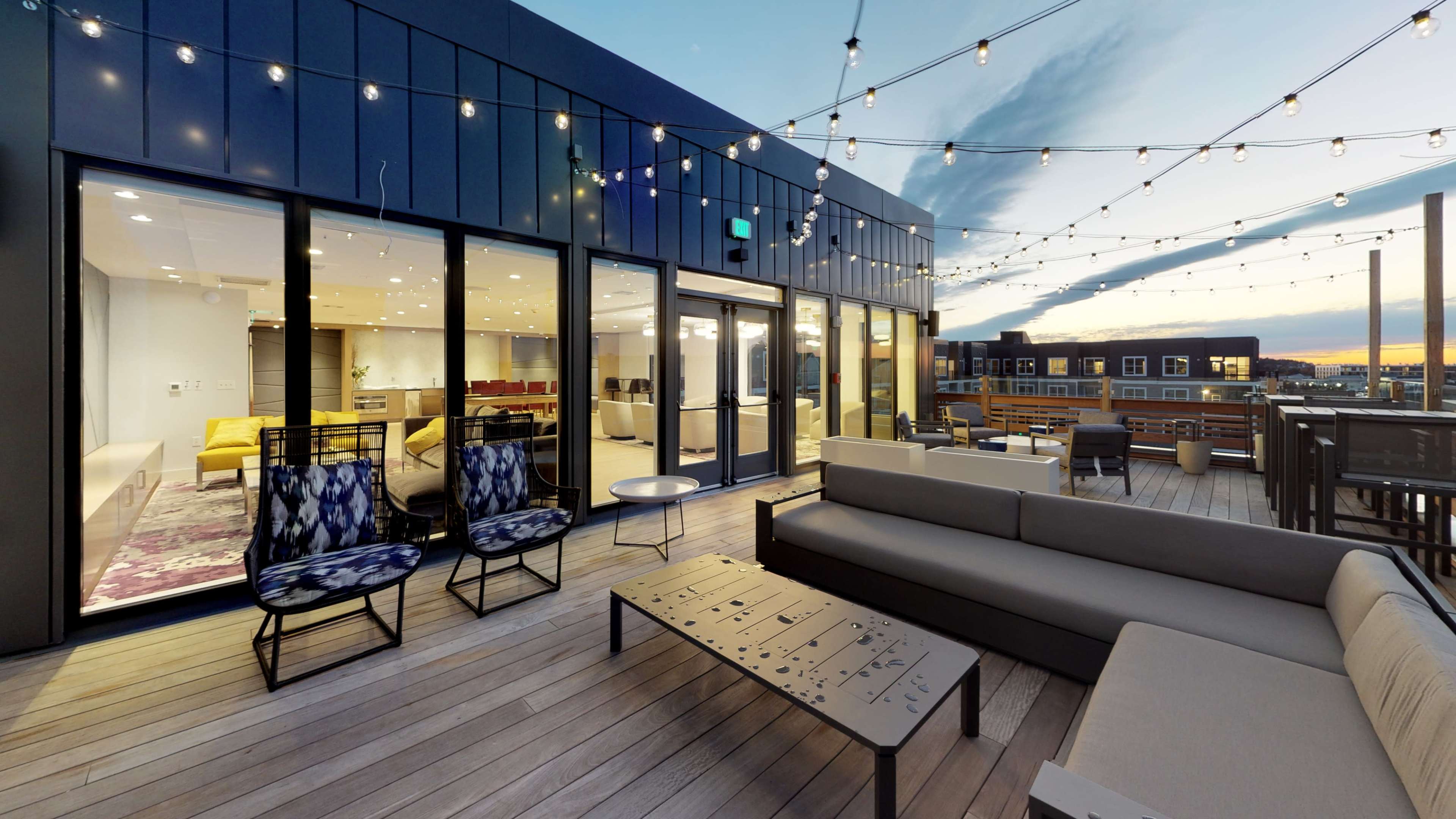 The rooftop terrace features modern furniture, string lights overhead, and large glass doors leading to an interior lounge area.