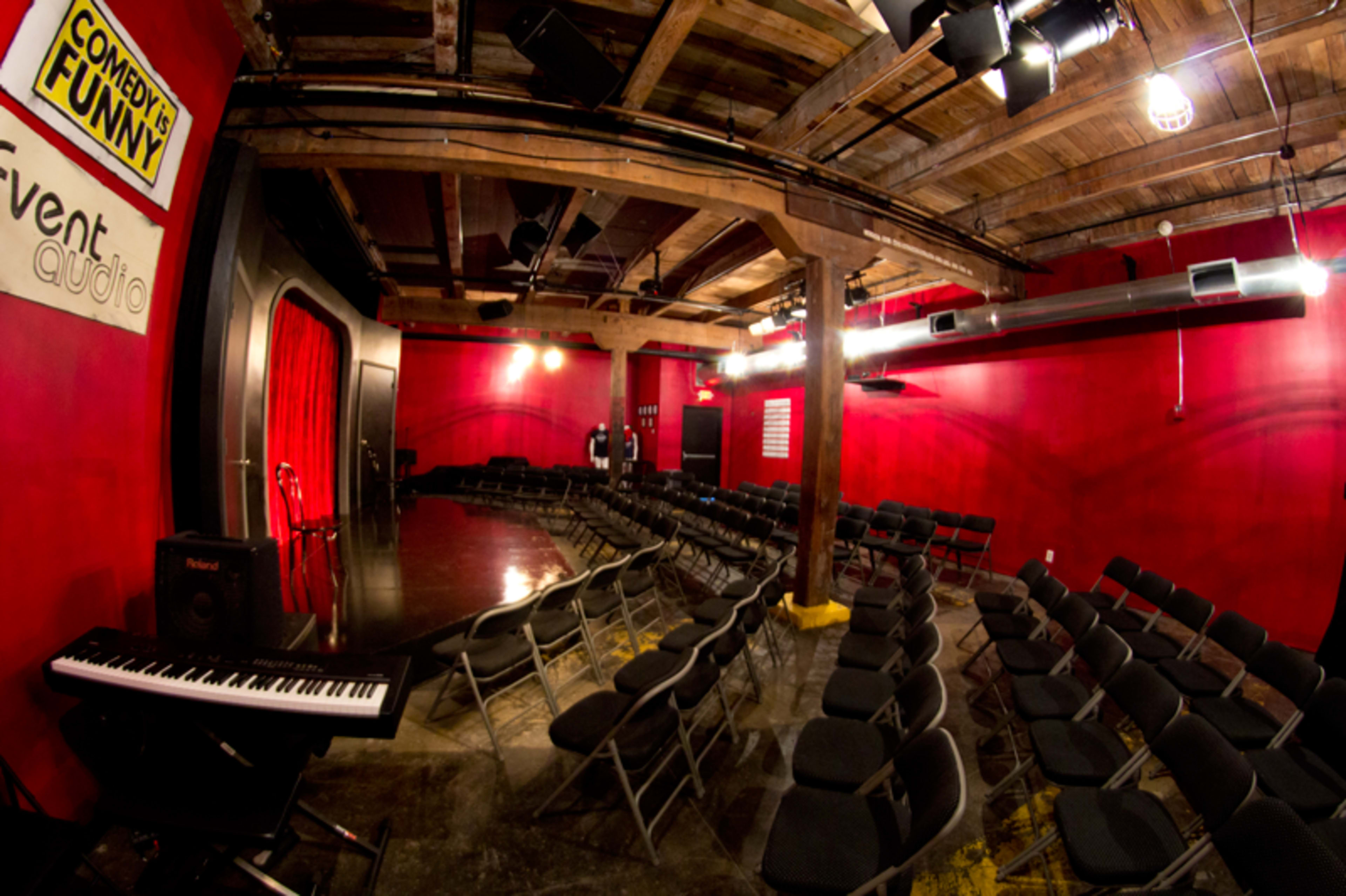 An empty comedy club with rows of black chairs facing a small stage adorned with a red curtain and a piano on the side.