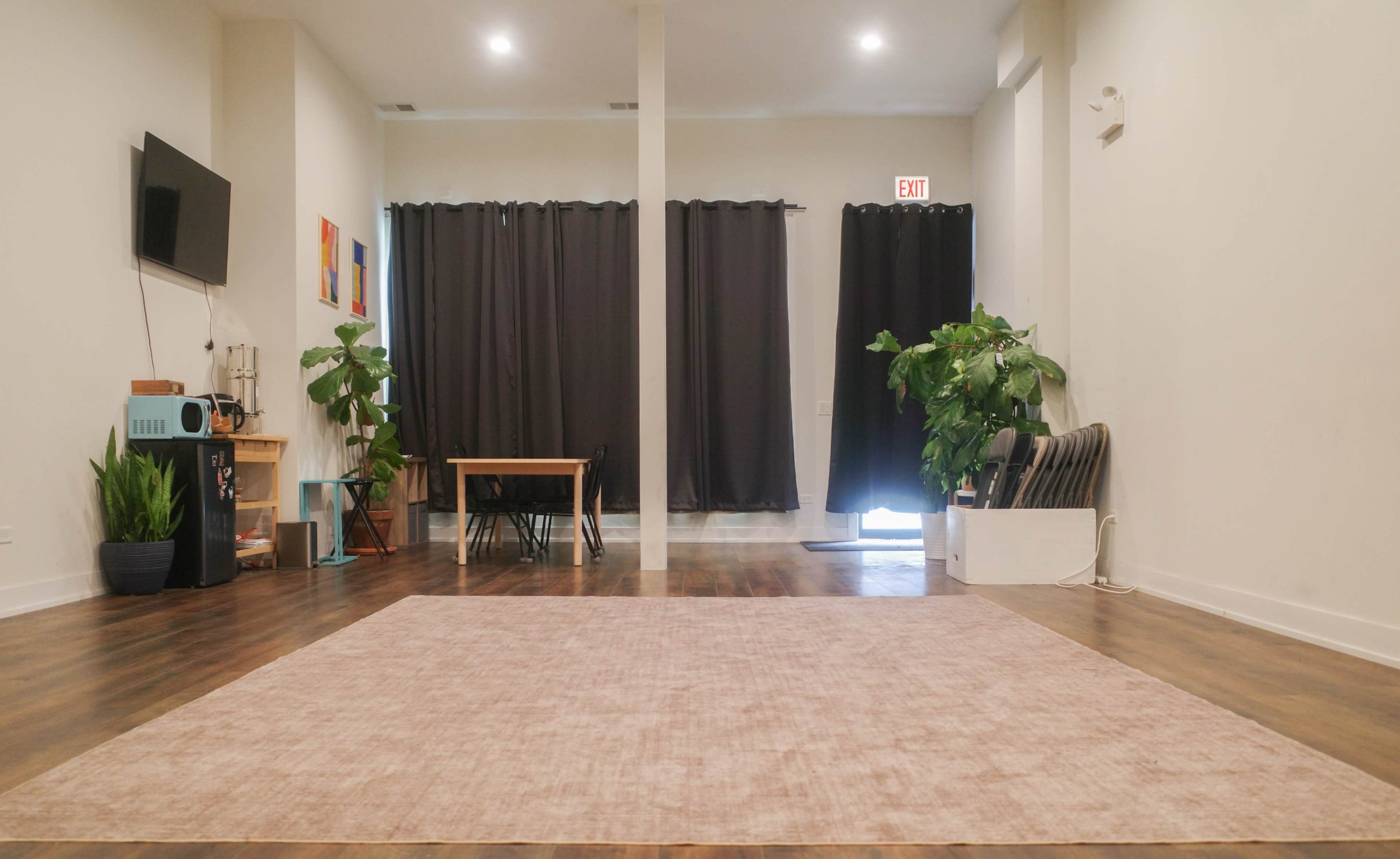 The image shows a spacious room with wooden floors, large curtains, a table at the back, and plants along the walls.