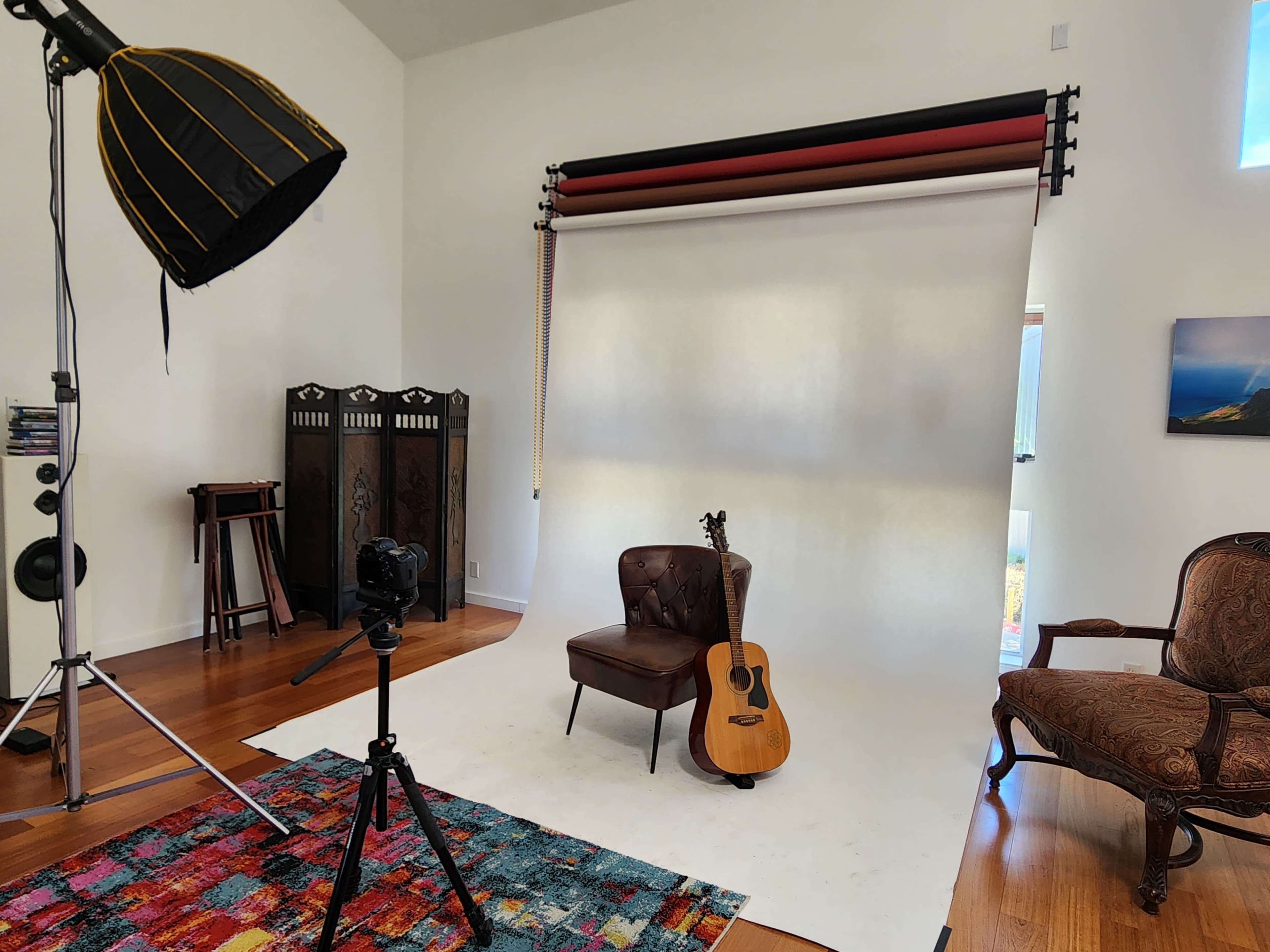 A photography studio setup with a brown leather chair, an acoustic guitar, a tripods-mounted camera, and a large softbox light in a well-lit room featuring hardwood floors and white walls.