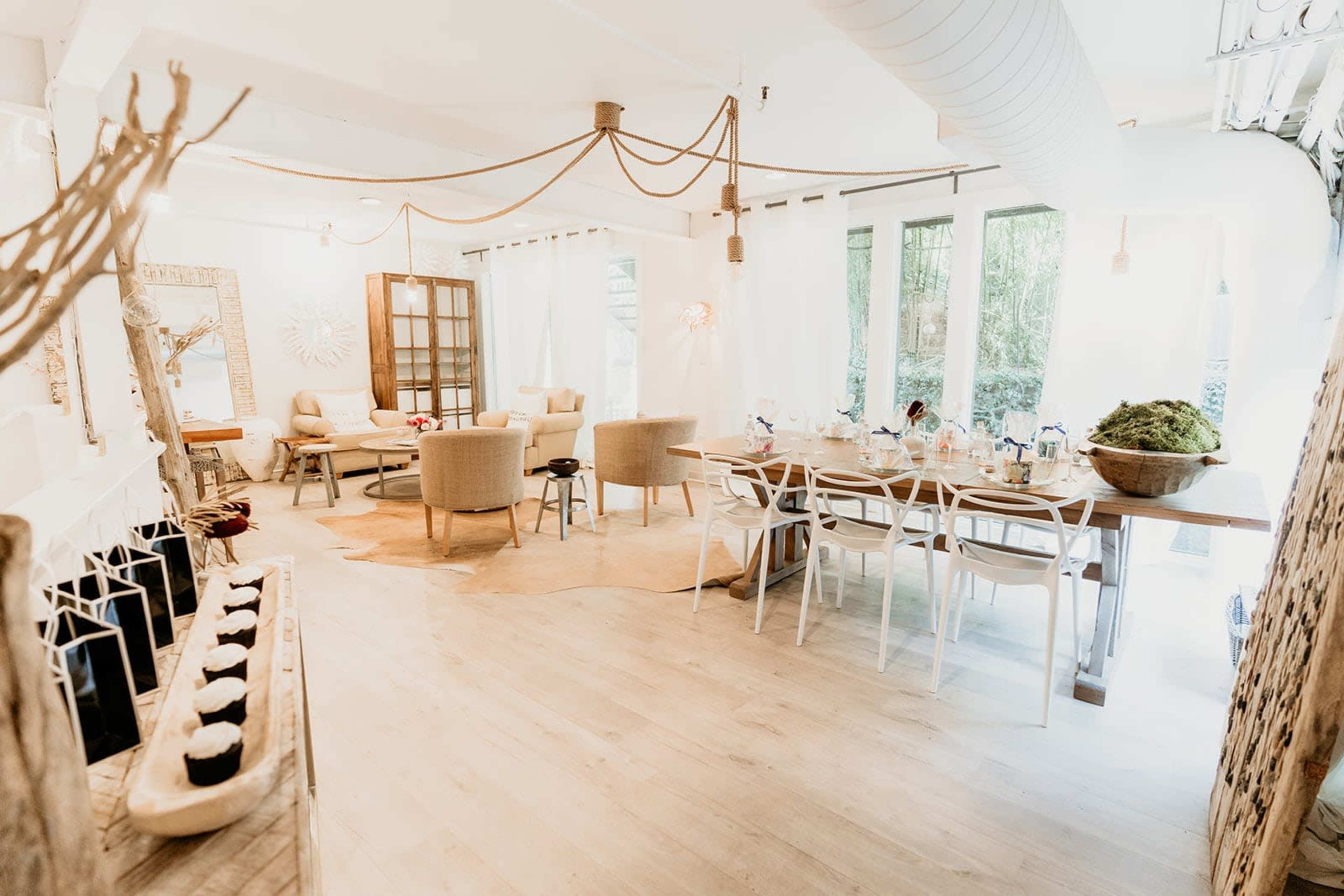 A spacious, well-lit room features a dining table, comfortable seating areas, and decorative elements, with large windows allowing natural light to enter.