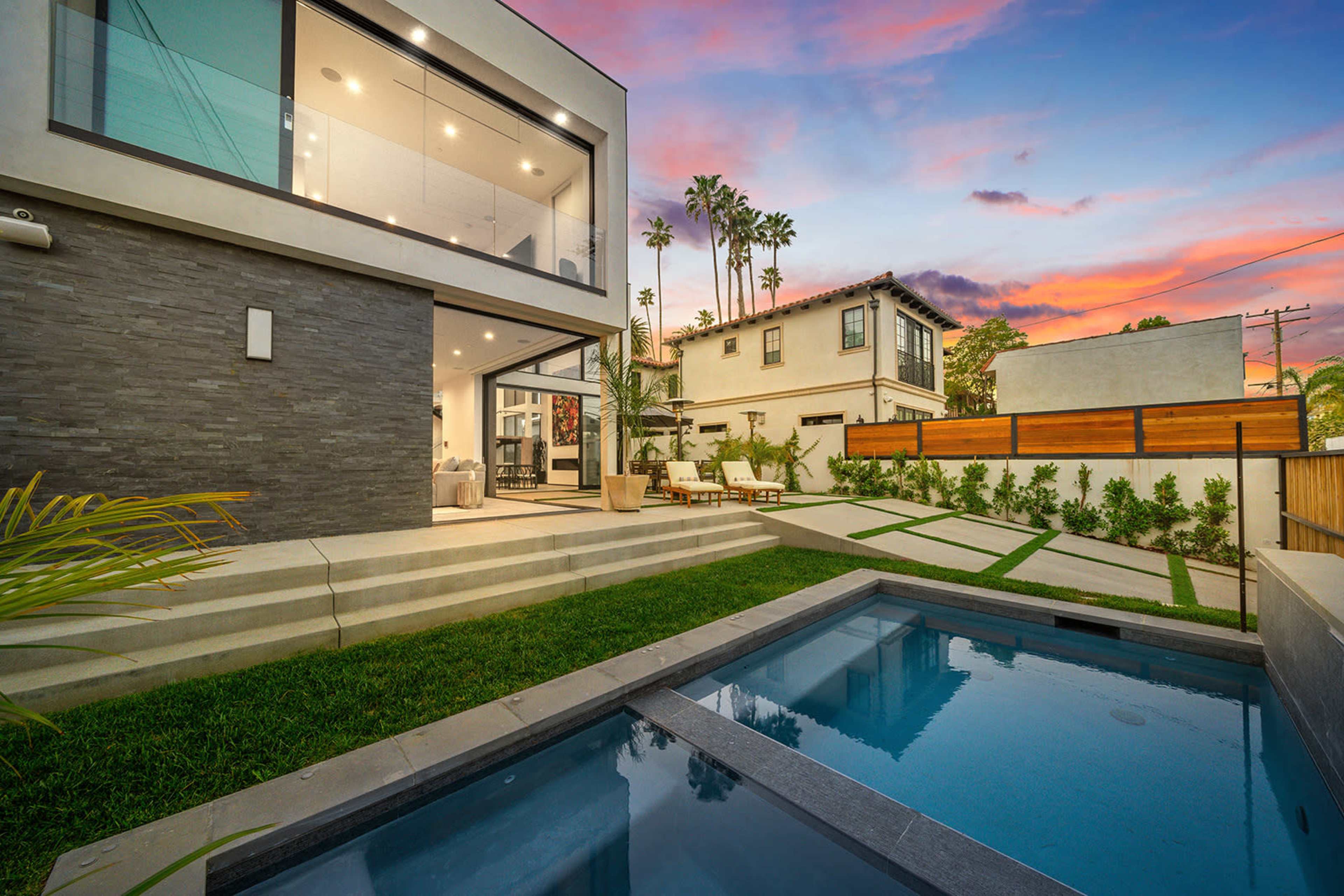 A modern house with large glass windows is surrounded by a neatly landscaped yard and a small swimming pool, set against a colorful sunset.