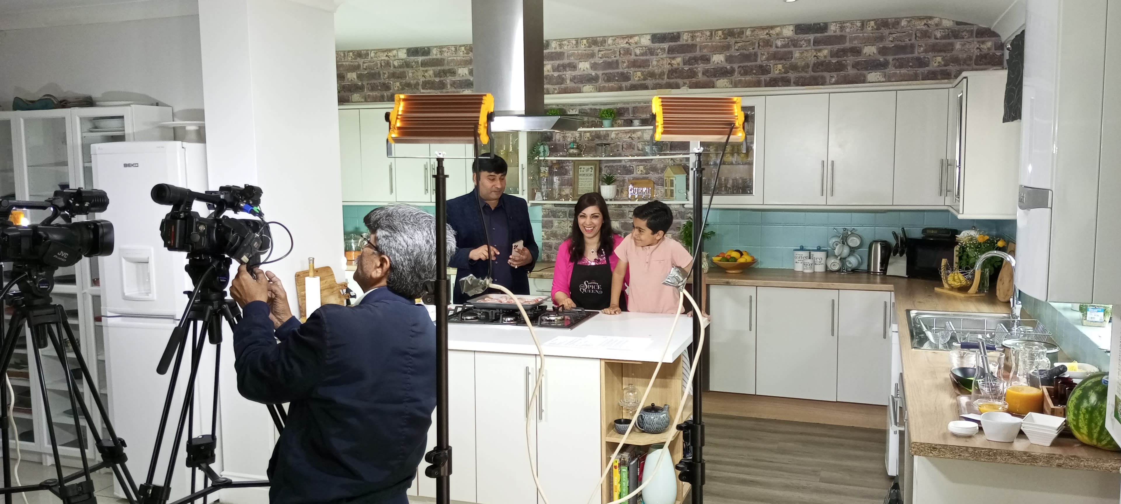 A cooking demonstration is taking place in a kitchen with cameras positioned to capture the scene as a host and two guests prepare food.