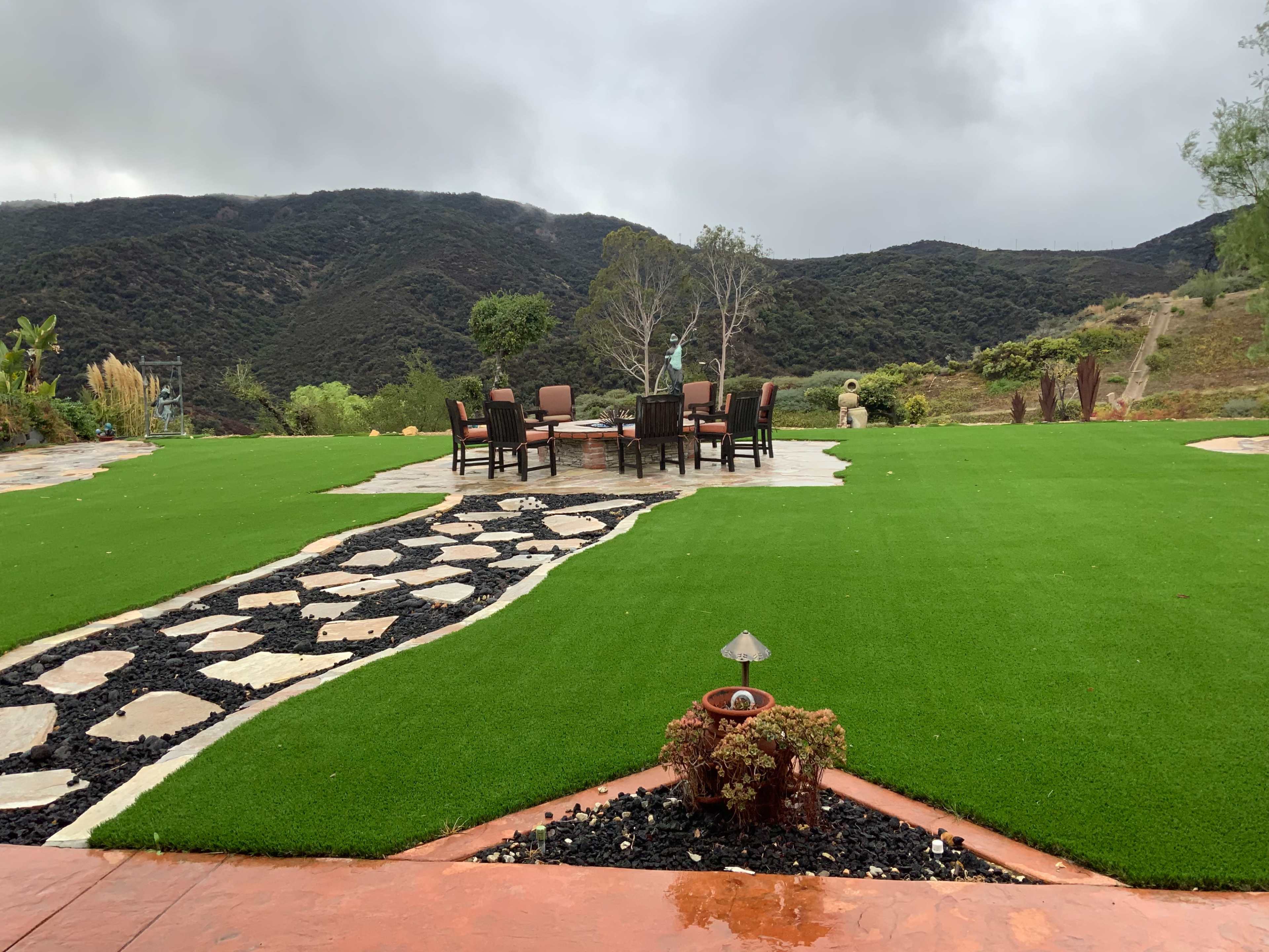 A patio with a stone pathway leads to a circular dining table surrounded by chairs on a green lawn, with mountains in the background under overcast skies.