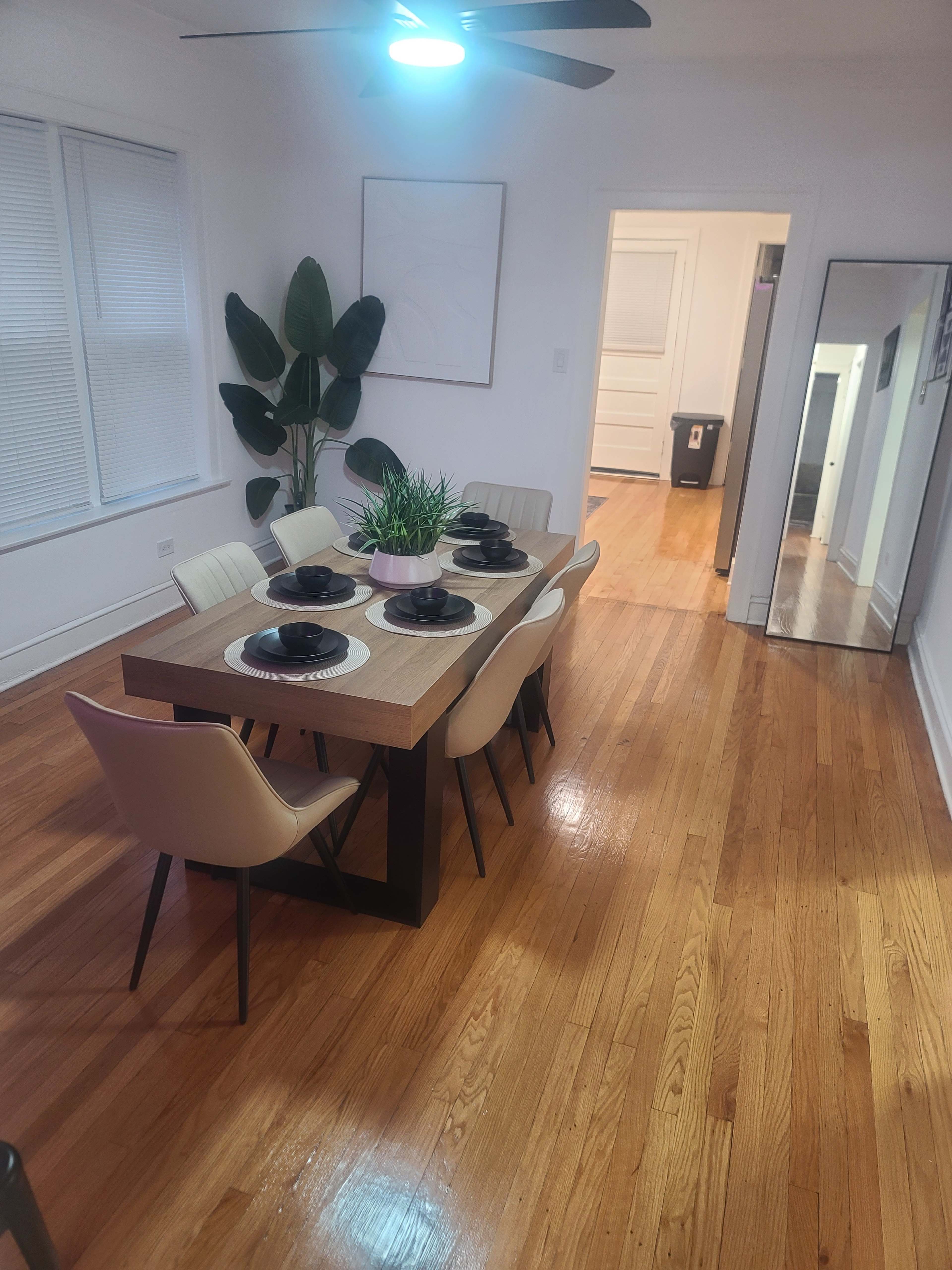 A dining room features a wooden table set with black plates and surrounded by light-colored chairs, with a large potted plant in the corner and a mirror on one wall.