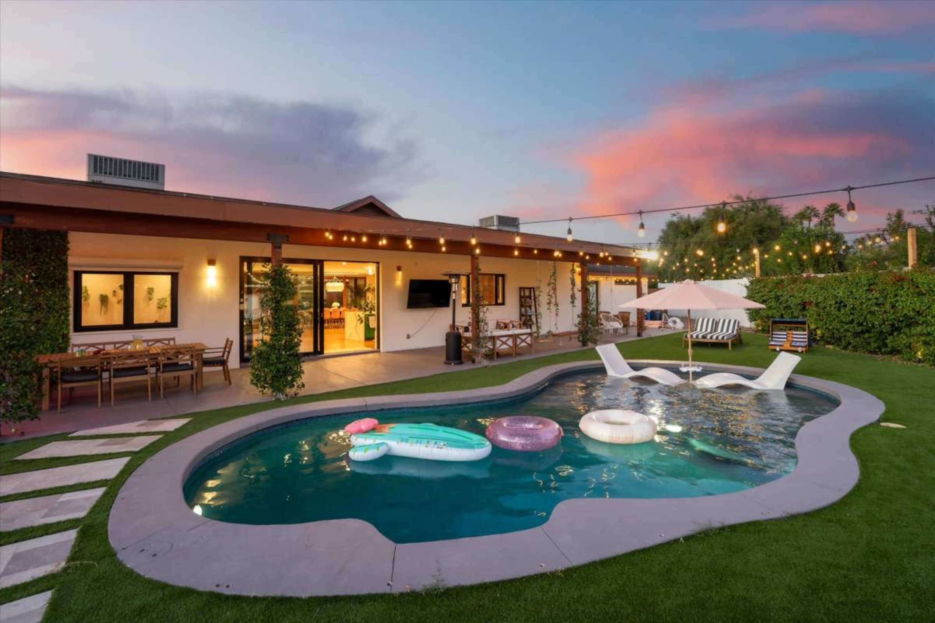 The image shows a modern backyard with a swimming pool, inflatable pool floats, outdoor seating, and string lights against a sunset sky.