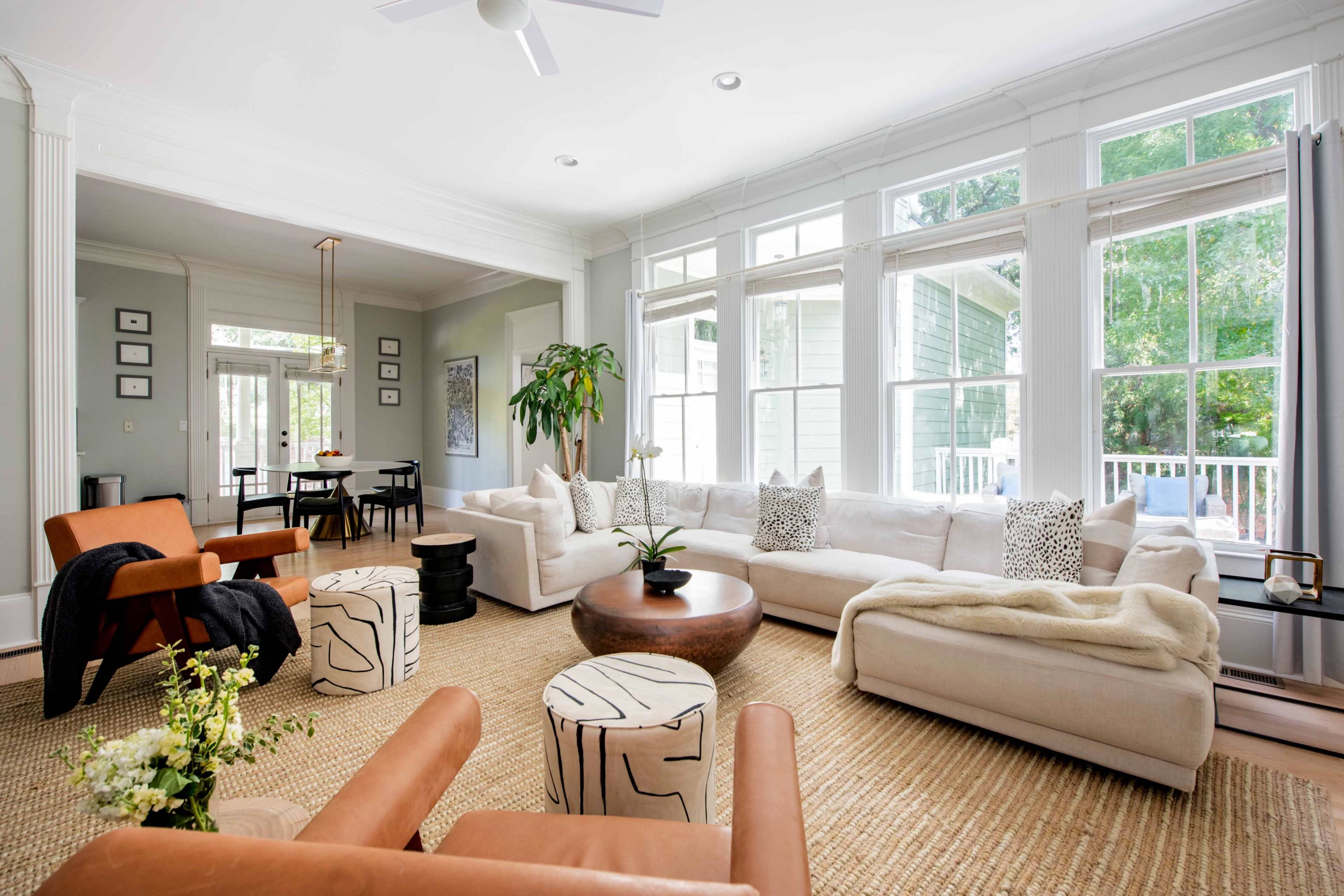 The living room features a white sectional sofa, a round wooden coffee table, and light-colored armchairs, with large windows providing natural light and a view of greenery outside.