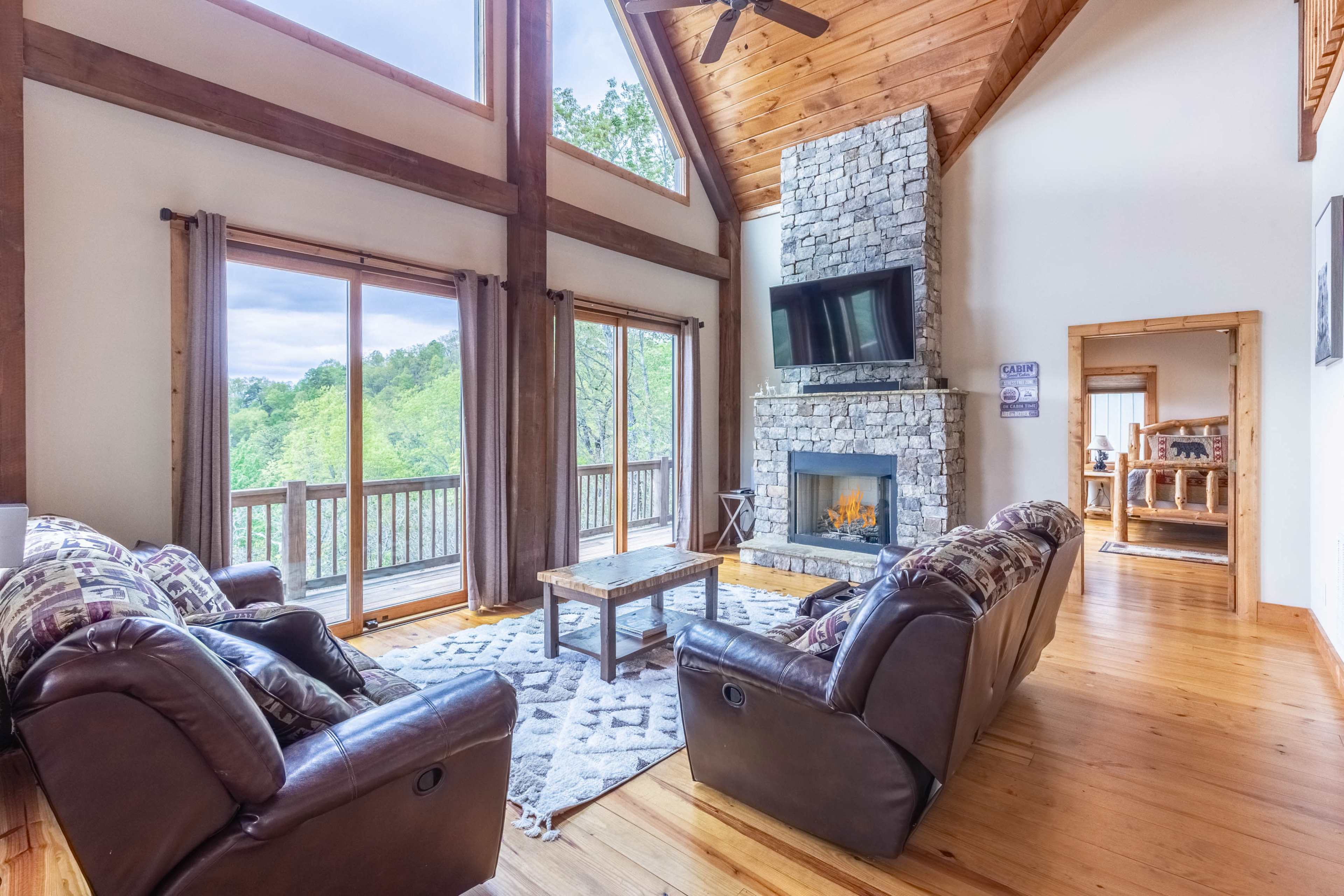 A spacious living room features a stone fireplace, large windows overlooking green scenery, and comfortable seating arranged around a wooden coffee table.
