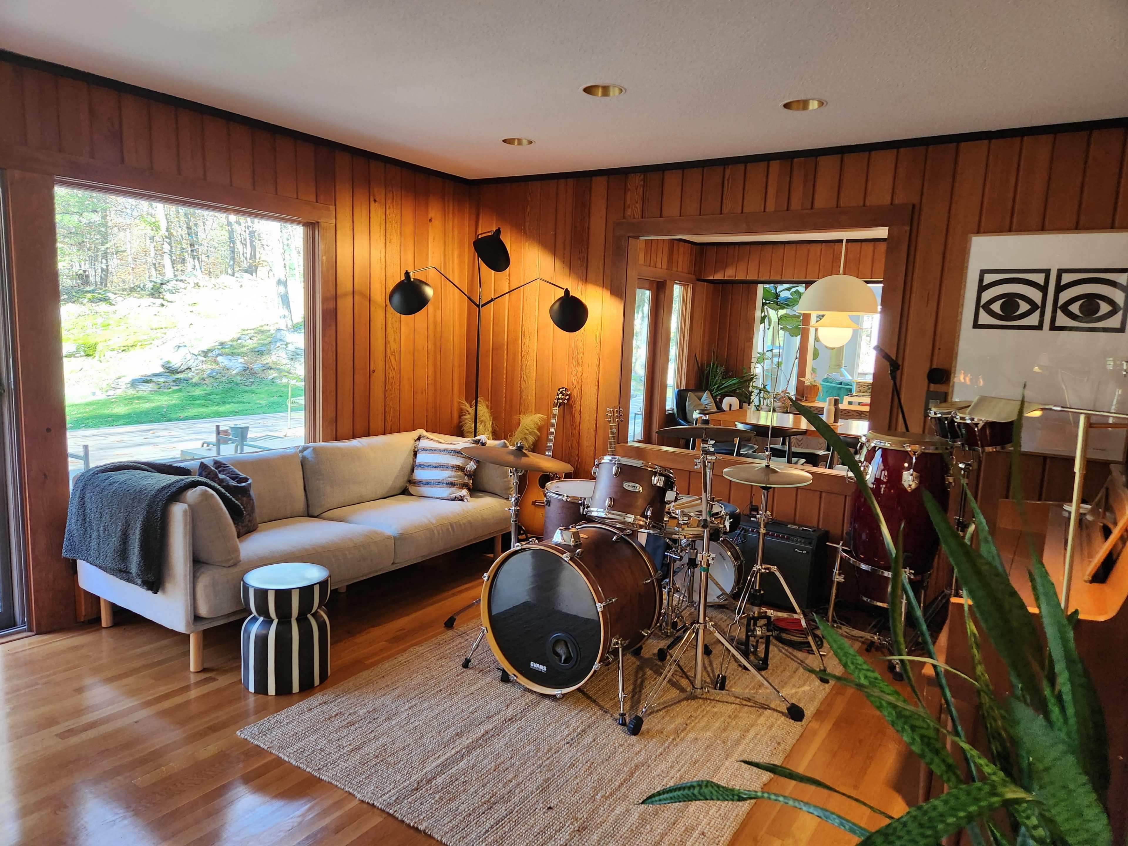 The image shows a living room with wooden walls, a couch, a coffee table, a drum set, and a large window that overlooks a garden.
