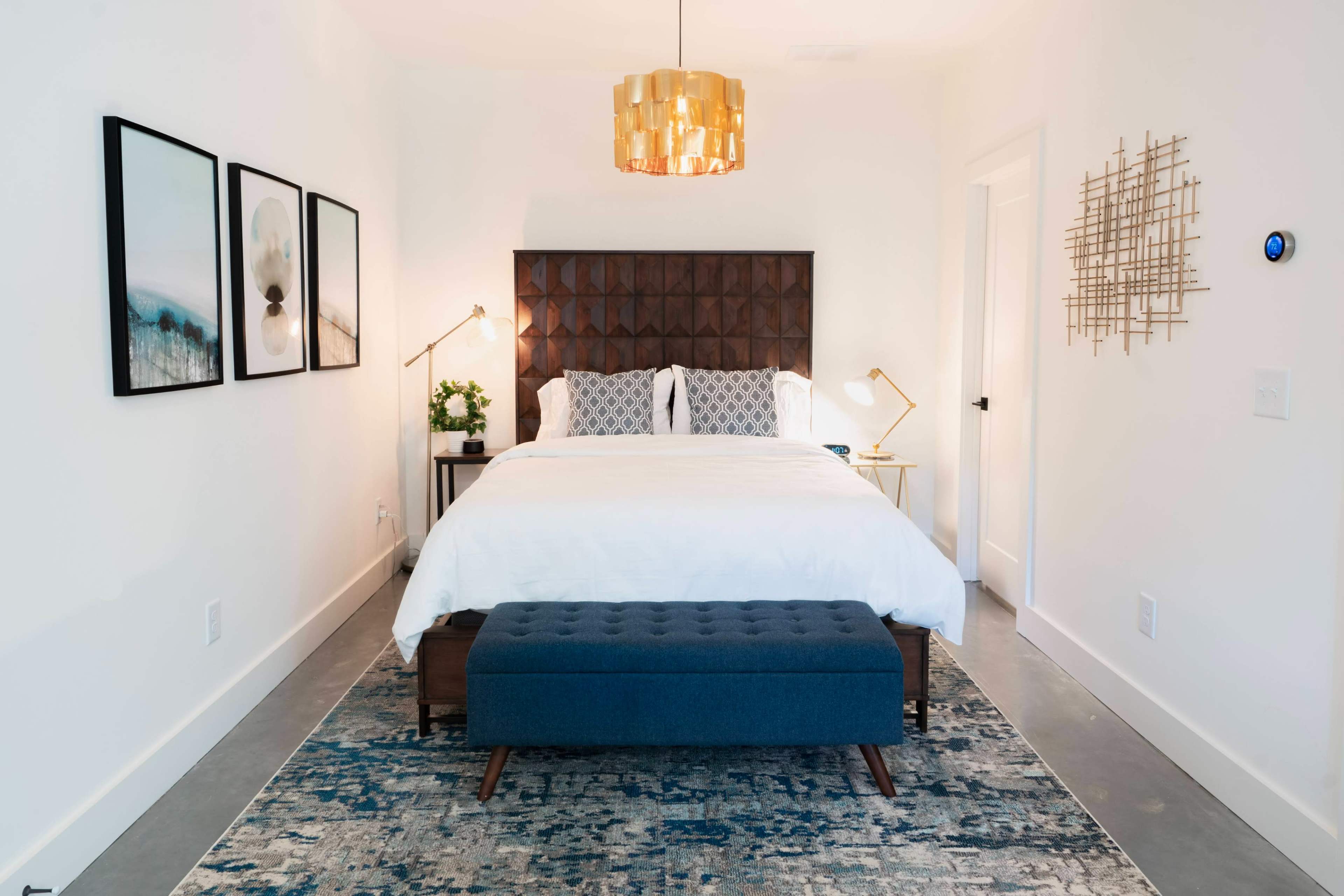 A modern bedroom features a large bed with a dark tufted headboard, flanked by two lamps, a decorative wall art arrangement, and a blue upholstered bench in front of the bed.