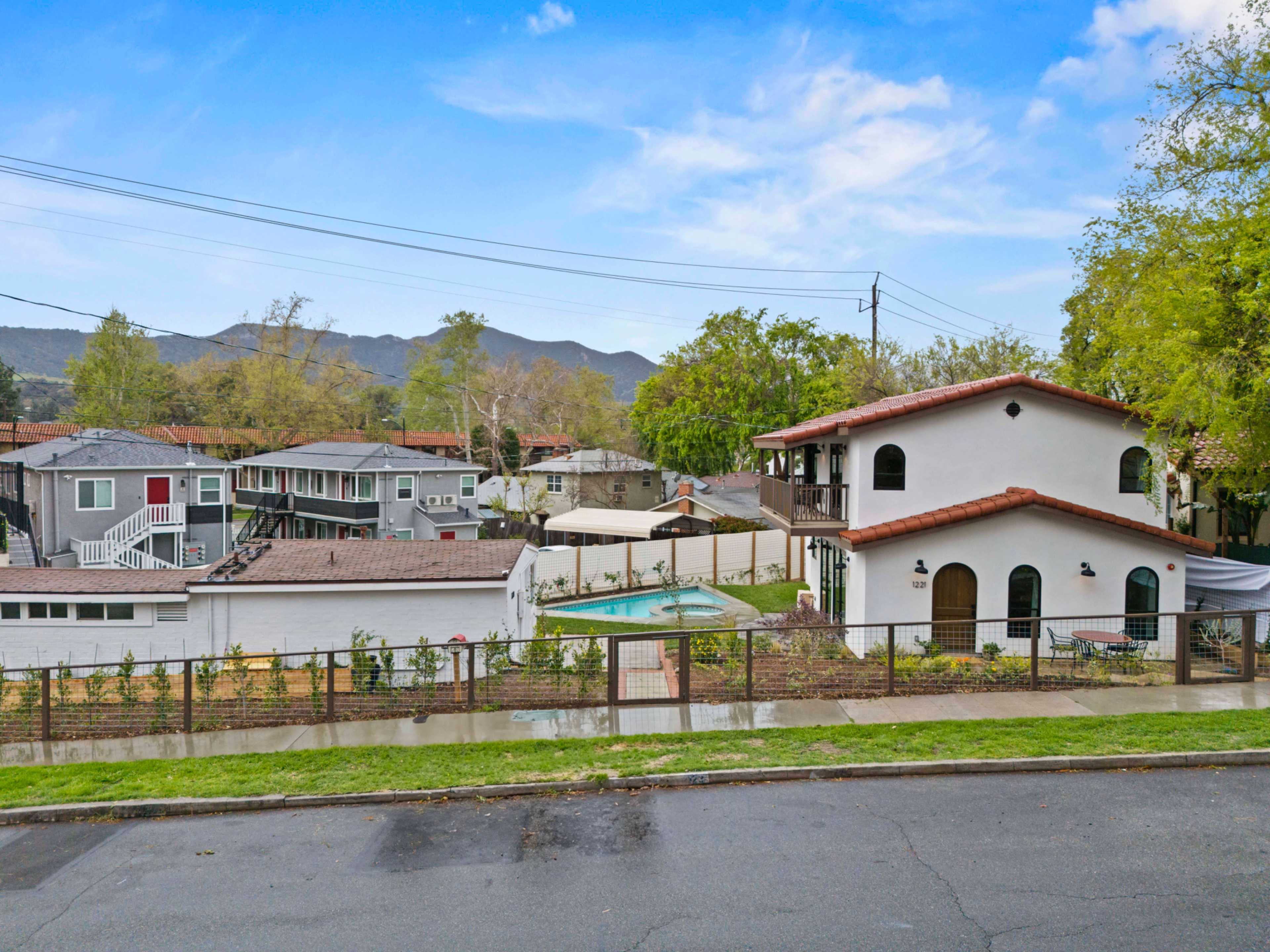 The image shows a suburban neighborhood with a mixture of single and multi-story homes, a fenced yard, and a small pool, set against a backdrop of hills.