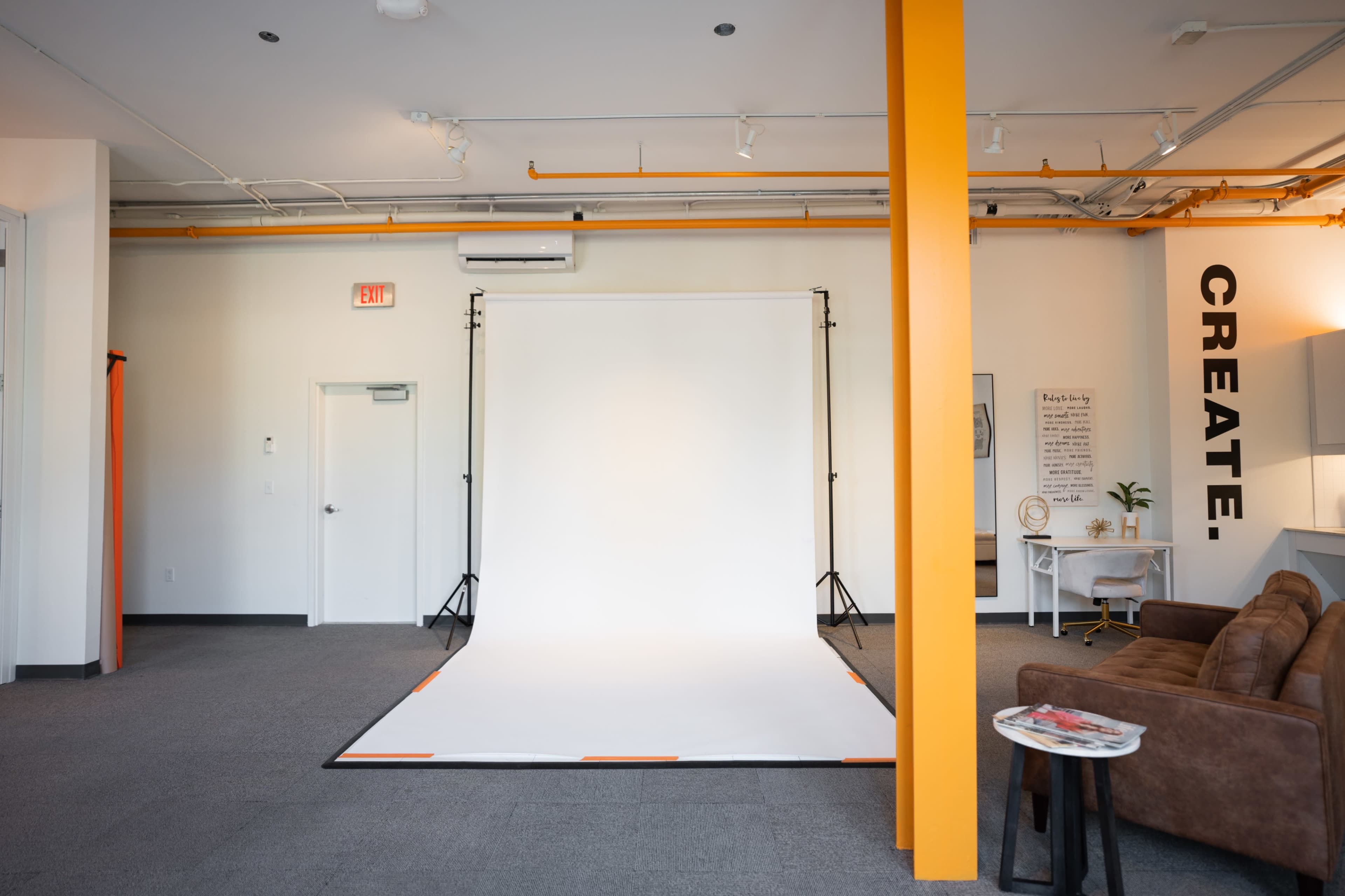 A spacious studio with a white backdrop set up for photography, featuring an orange accent wall, a brown sofa, and artwork on the walls.