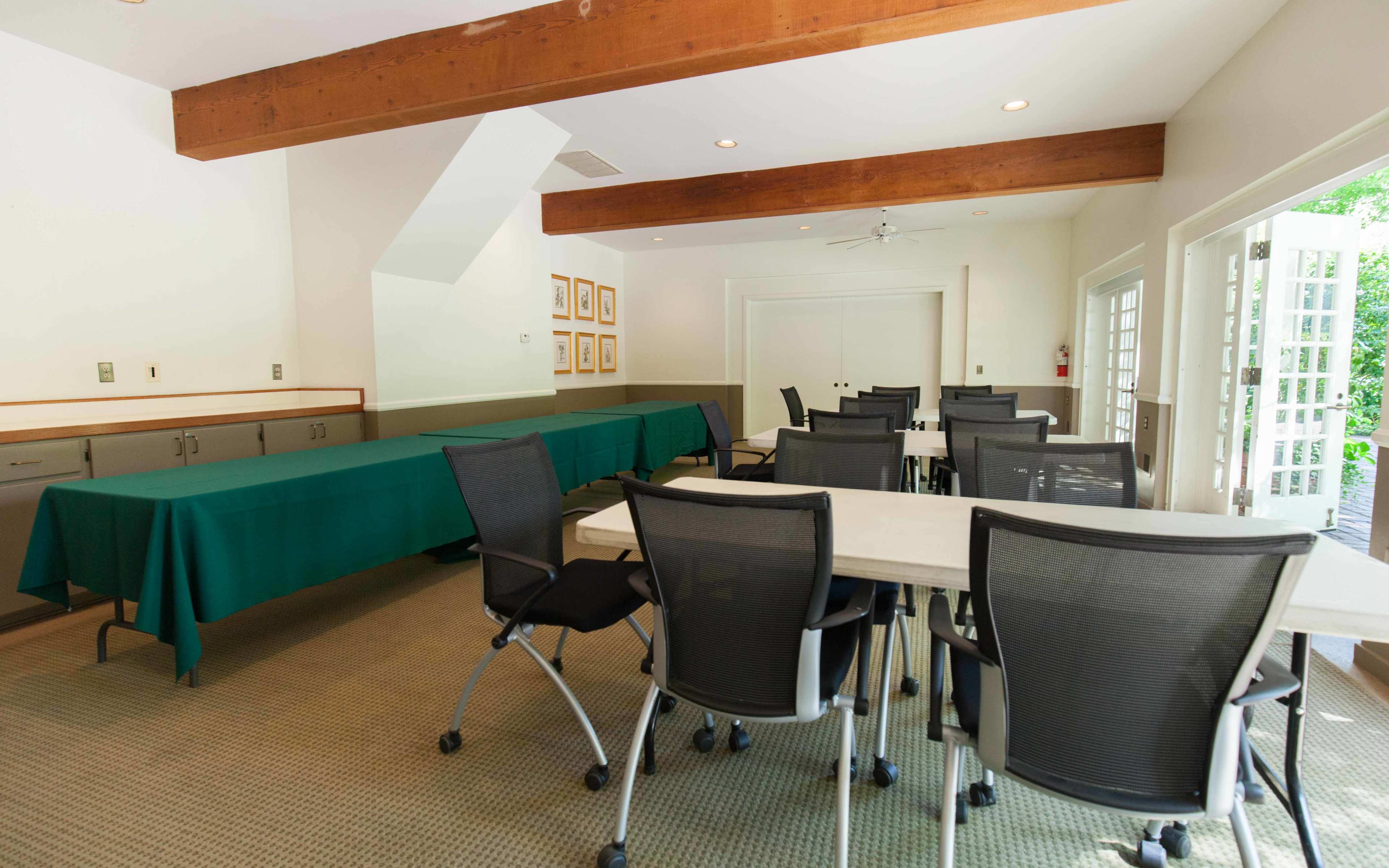 A conference room features a rectangular table covered with a green tablecloth, surrounded by several black rolling chairs, with large windows and a door leading outside.