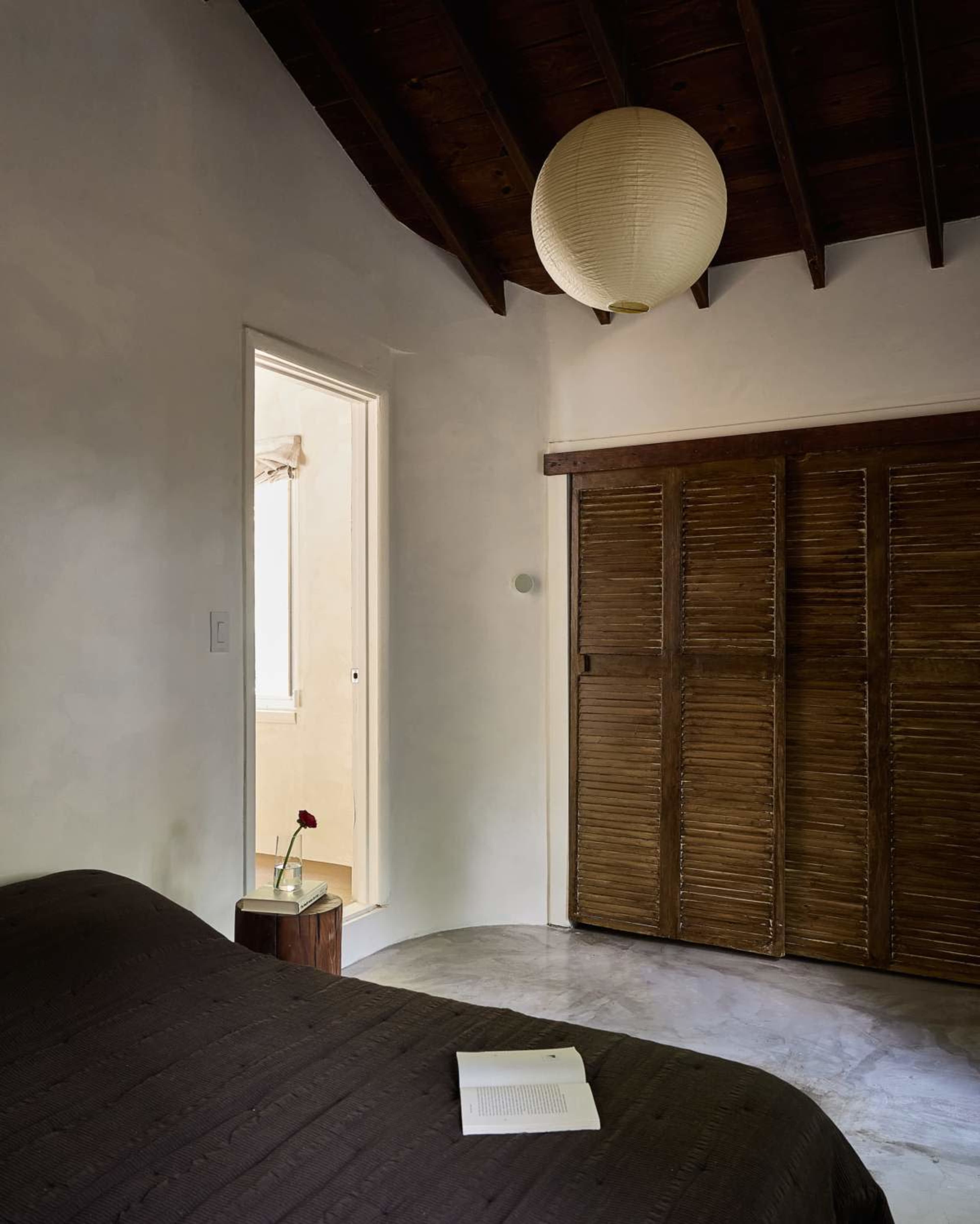 A dimly lit bedroom features a bed with a dark cover, a wooden cabinet with slatted doors, and a small table holding a single flower next to an open book.