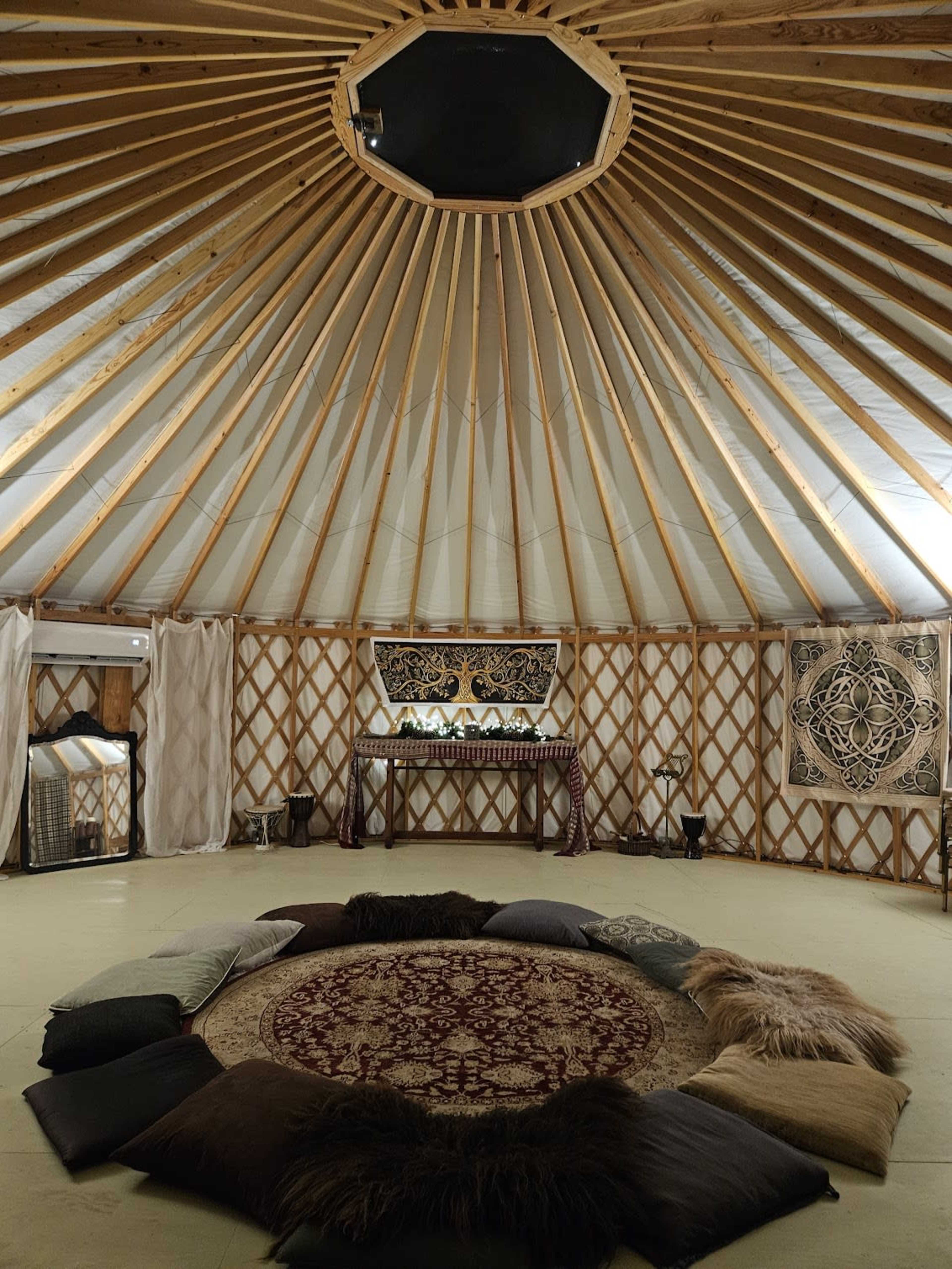 The interior of a yurt features a circular seating arrangement of cushions surrounding a central rug, with wooden beams supporting a circular skylight above.