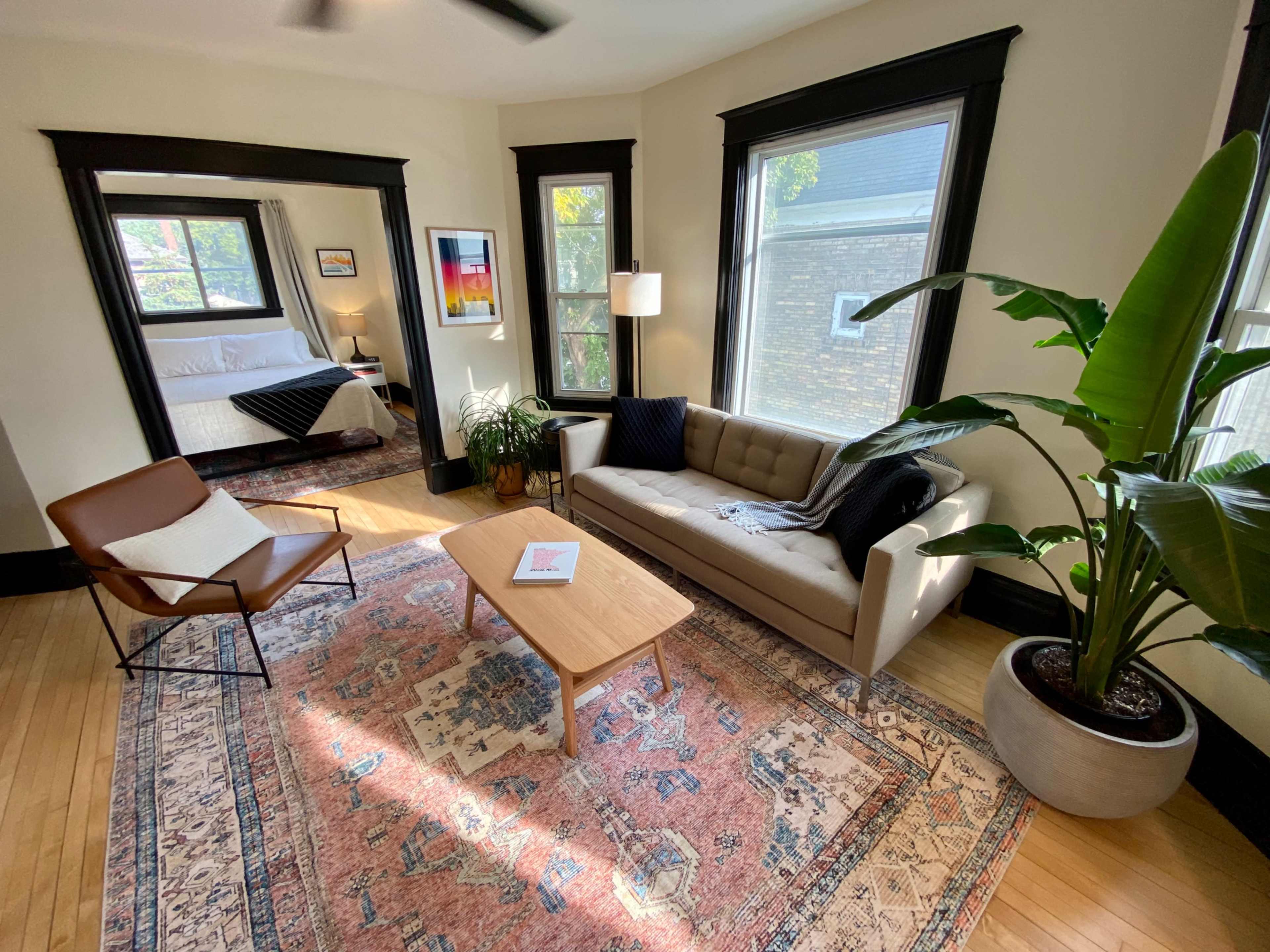 The image shows a living room with a sofa, a coffee table, a chair, a potted plant, and large windows that provide natural light.