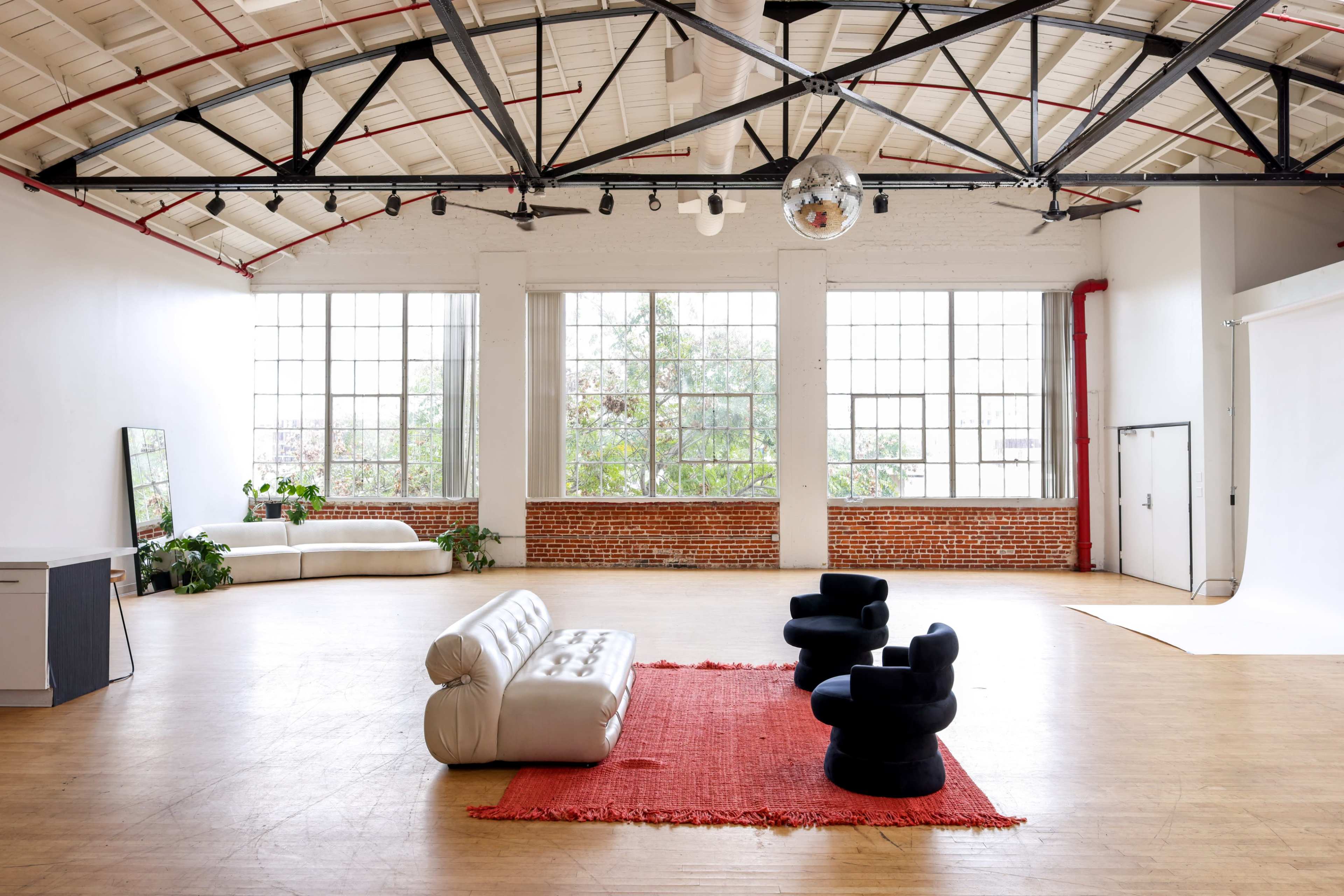 The image shows a spacious, well-lit loft with large windows, featuring a red area rug, a white sofa, and two black chairs.