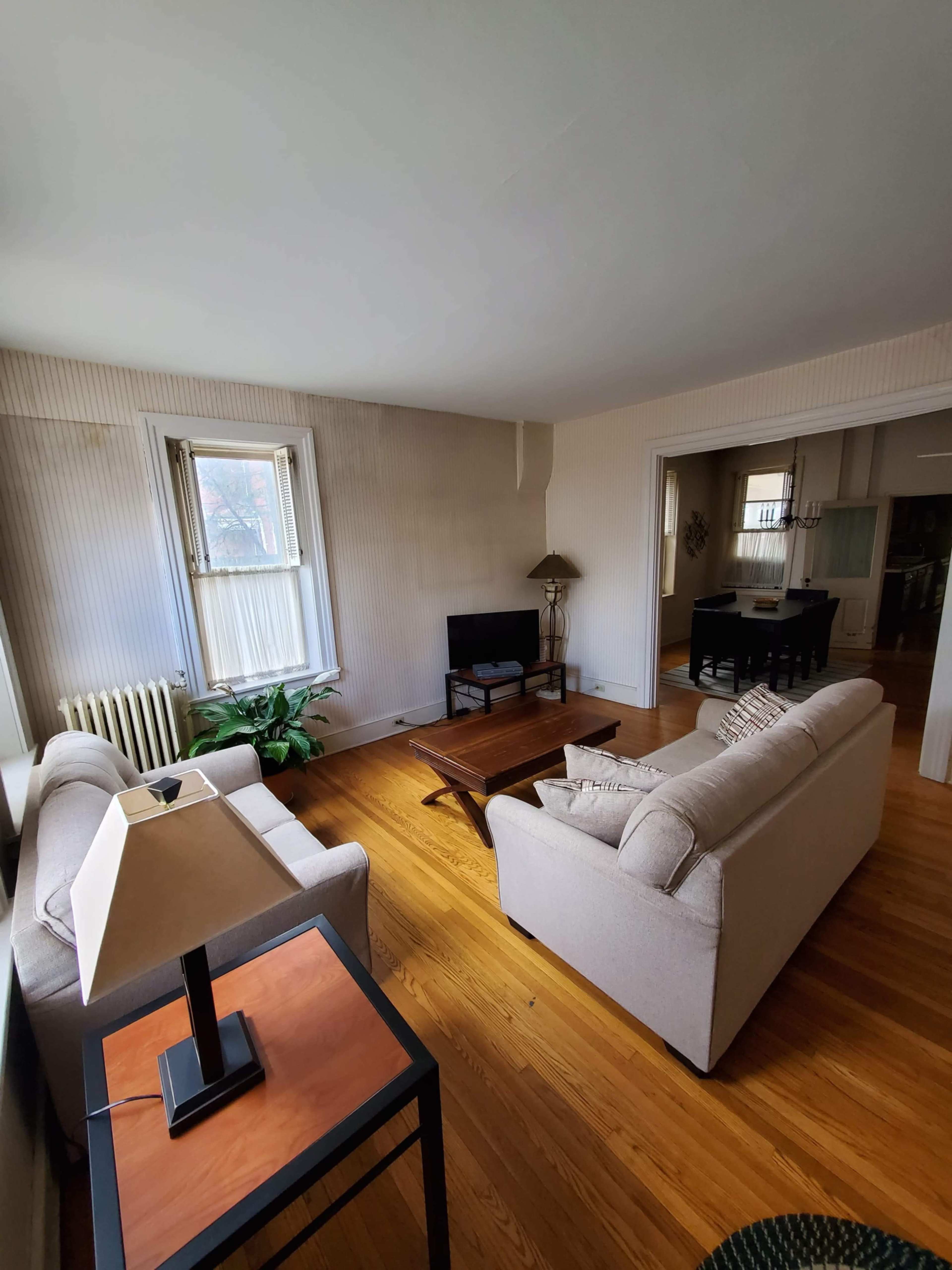 A living room with two sofas, a coffee table, a lamp, and a television, featuring hardwood floors and large windows.