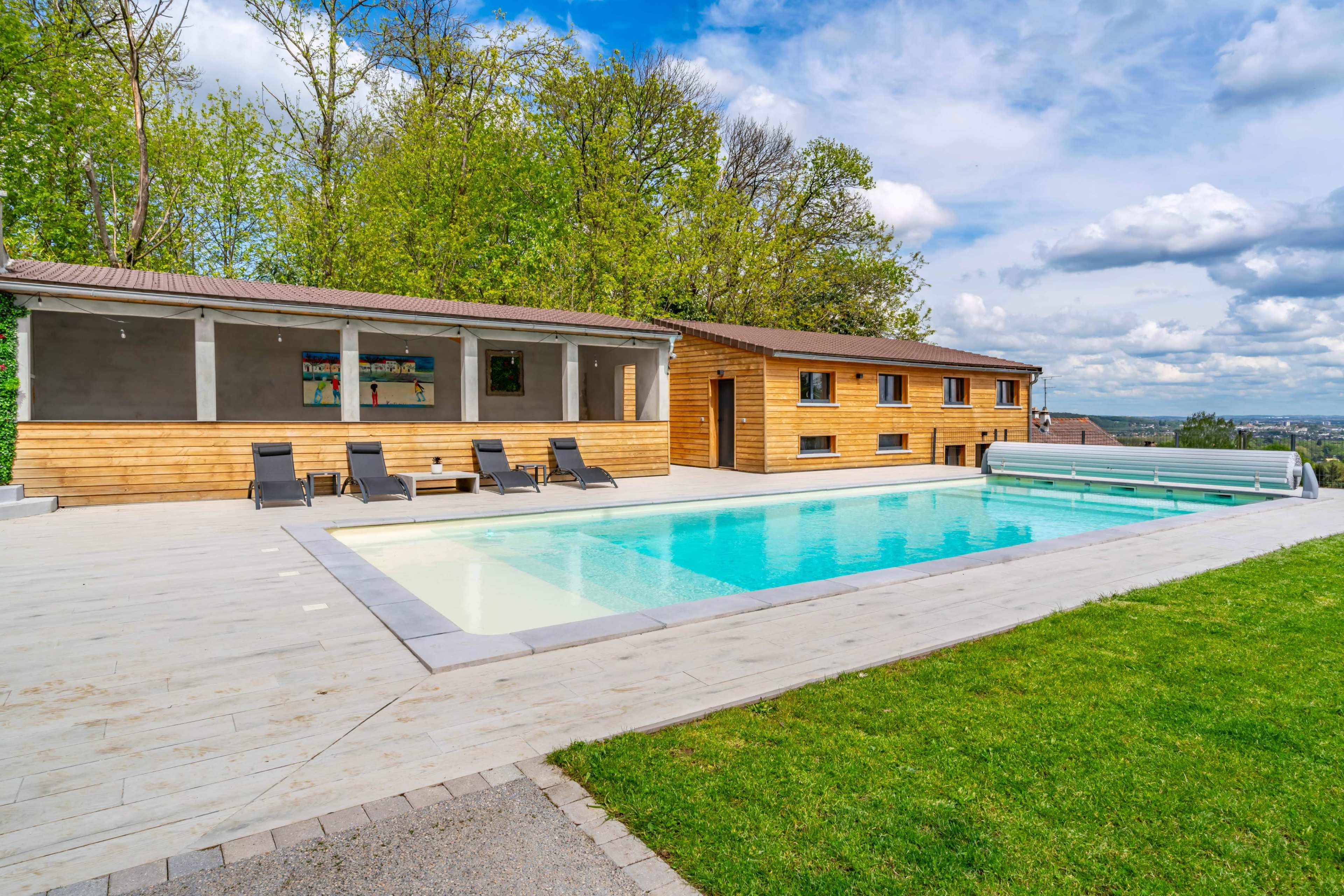 A modern wooden house with a sun terrace and a swimming pool surrounded by green grass and trees.
