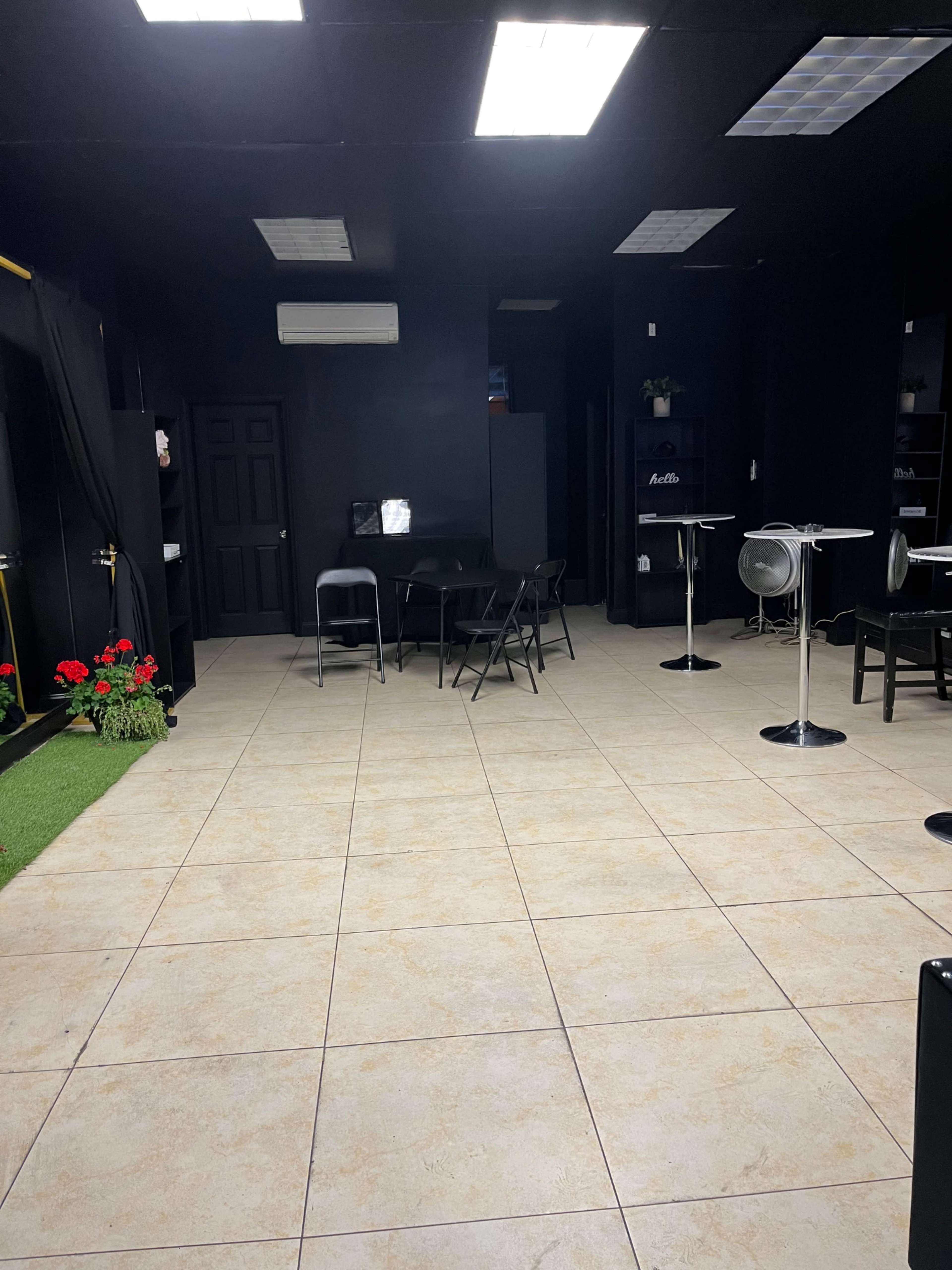 An empty room with black walls, tiled flooring, a few tables and chairs, and potted plants near the entrance.