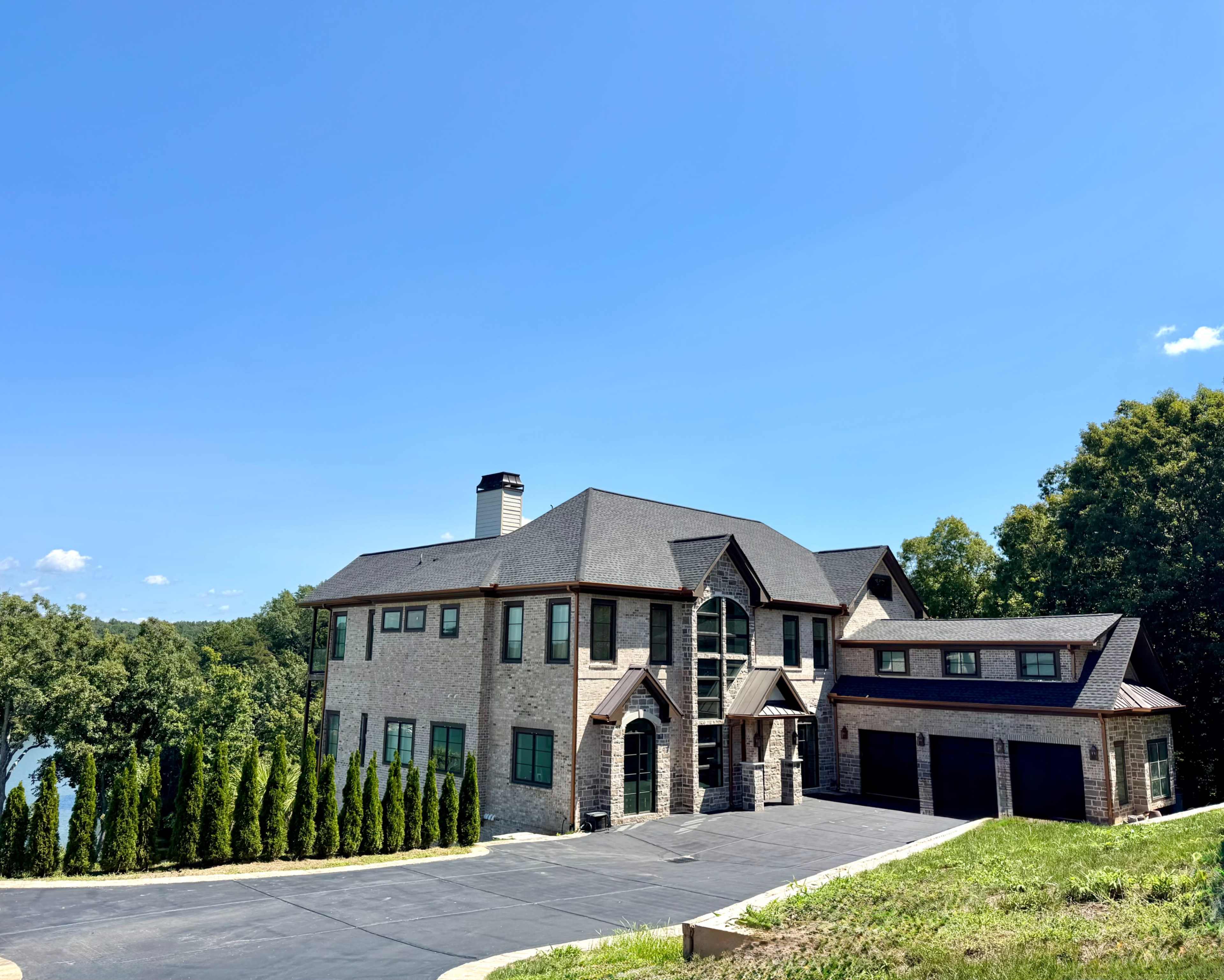 A large, stone-faced house with multiple windows and a three-car garage is situated on a green hillside beside a lake.