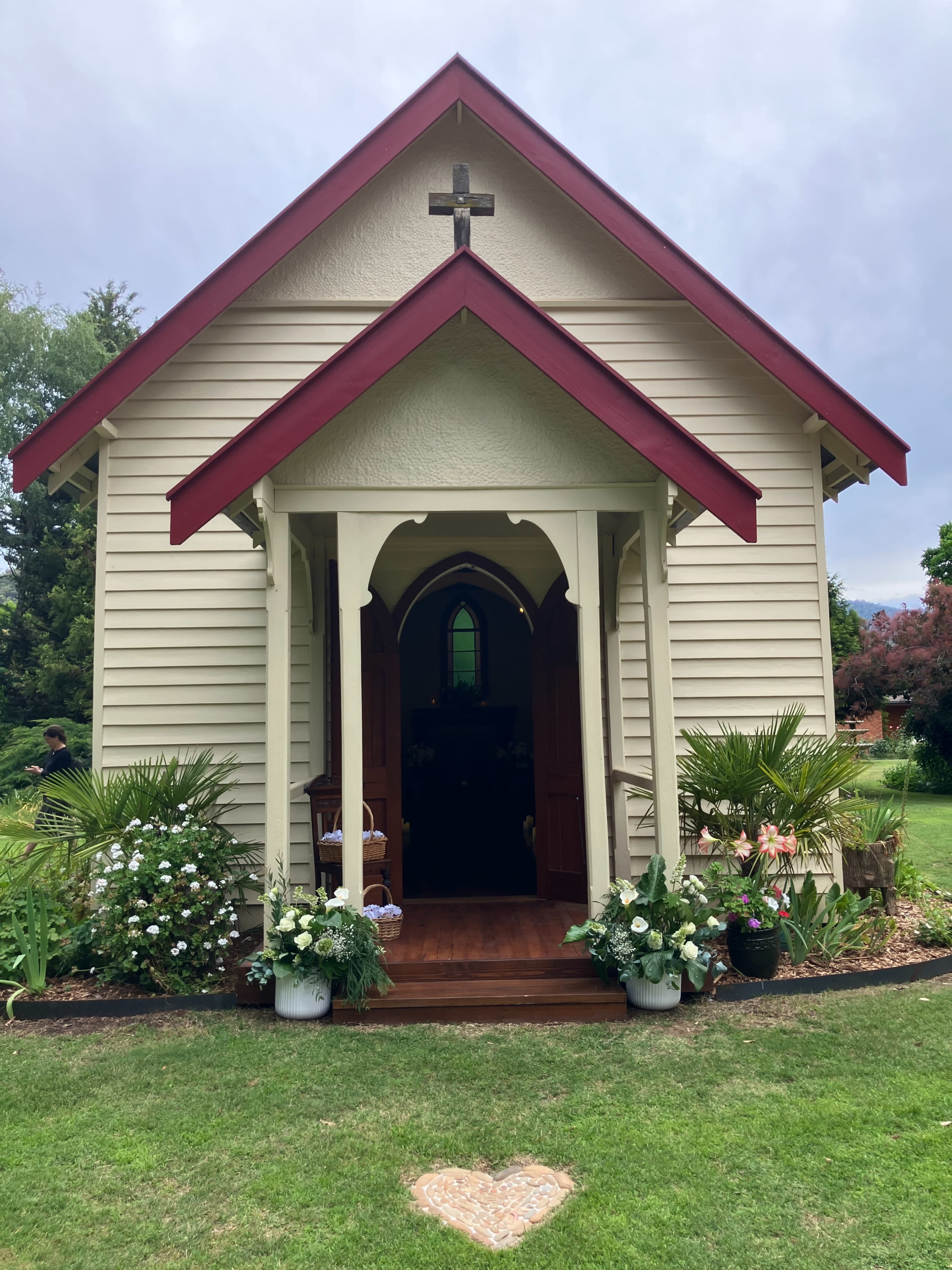 A small, quaint church with a red roof and cross stands surrounded by flower beds and greenery.