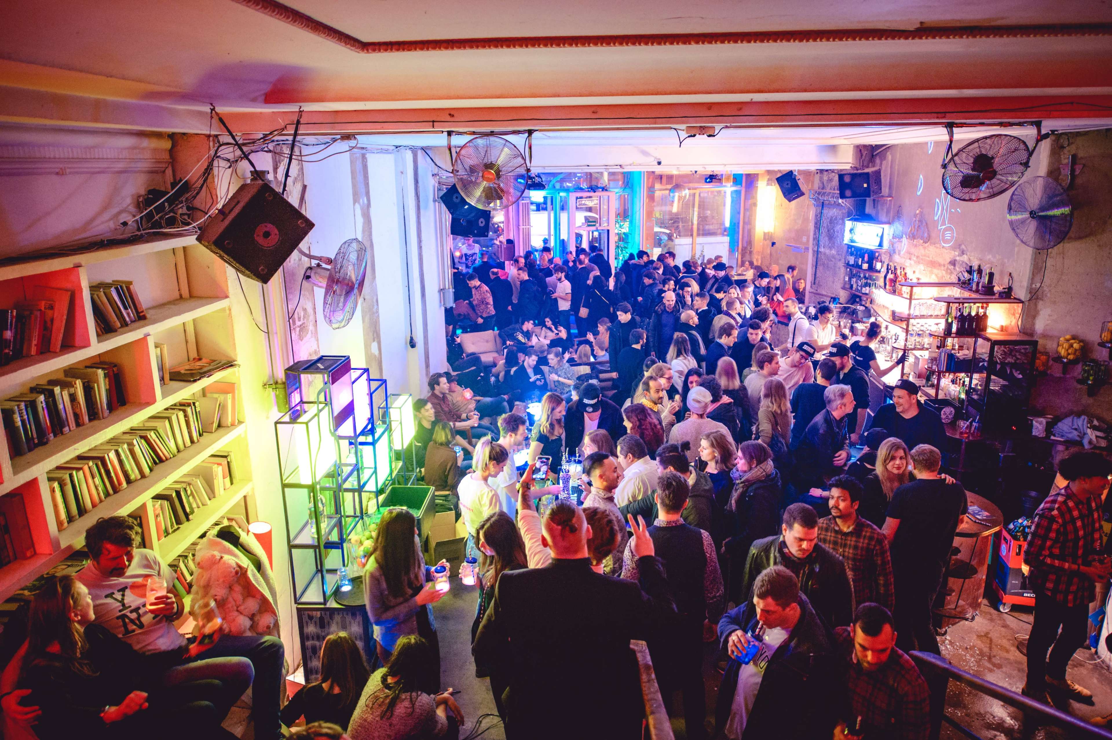 A lively crowd fills a vibrant bar or club, featuring colorful lighting and shelves stocked with books.