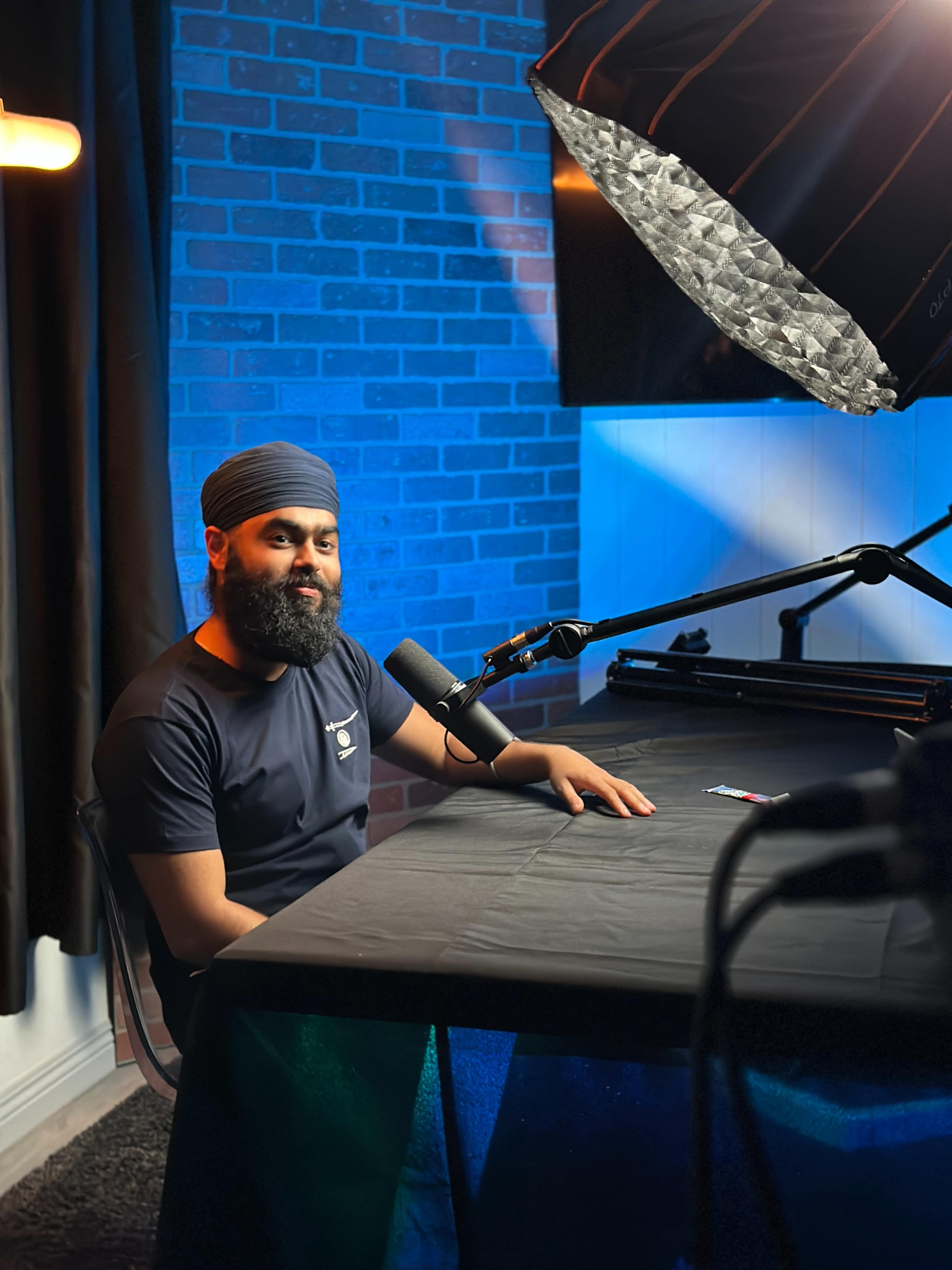 A man with a beard and a turban sits at a desk with a microphone in front of him, illuminated by soft lighting in a room with a brick wall.