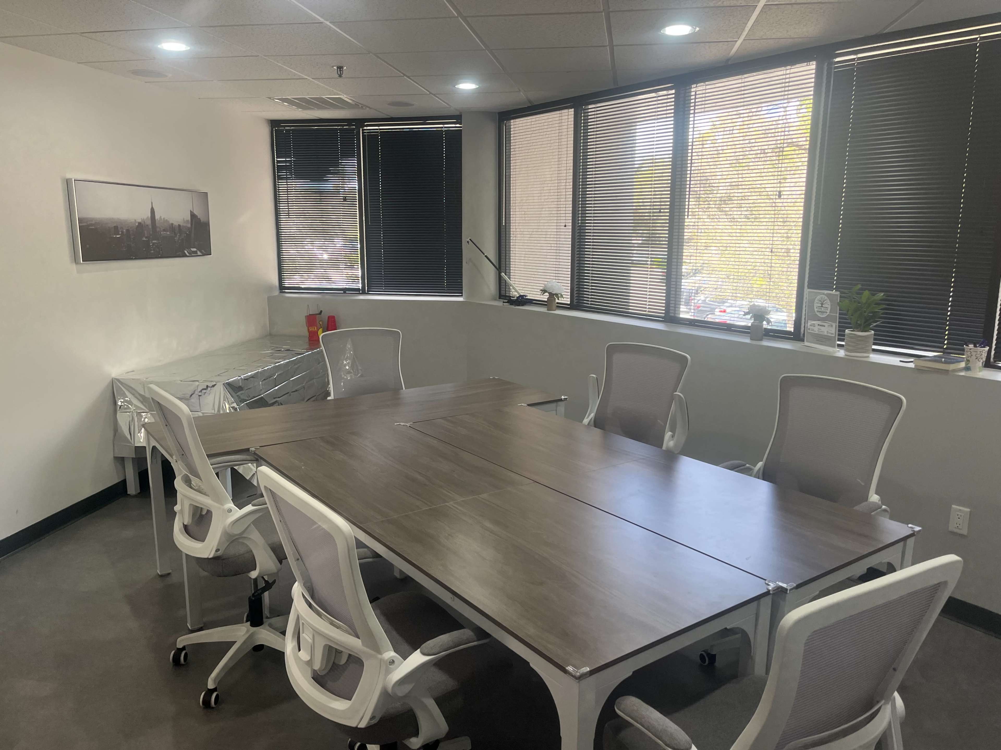 The image shows a conference room with a large rectangular table surrounded by six white chairs, windows with blinds, and a few decorative items on the walls and table.