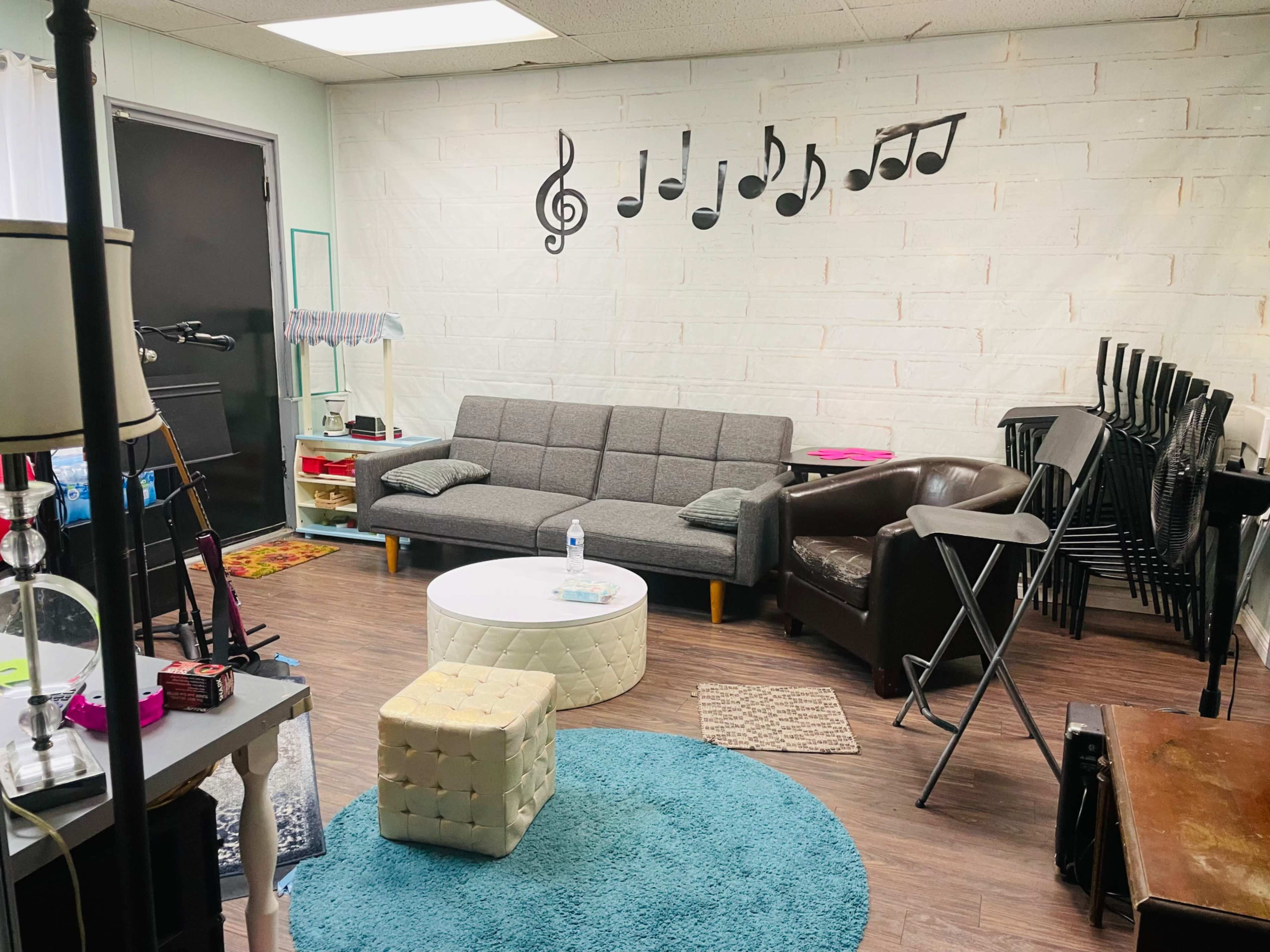 A room with a gray sofa, a brown armchair, a round coffee table, and music notes decor on a white wall, alongside various furniture and items stacked in the corner.
